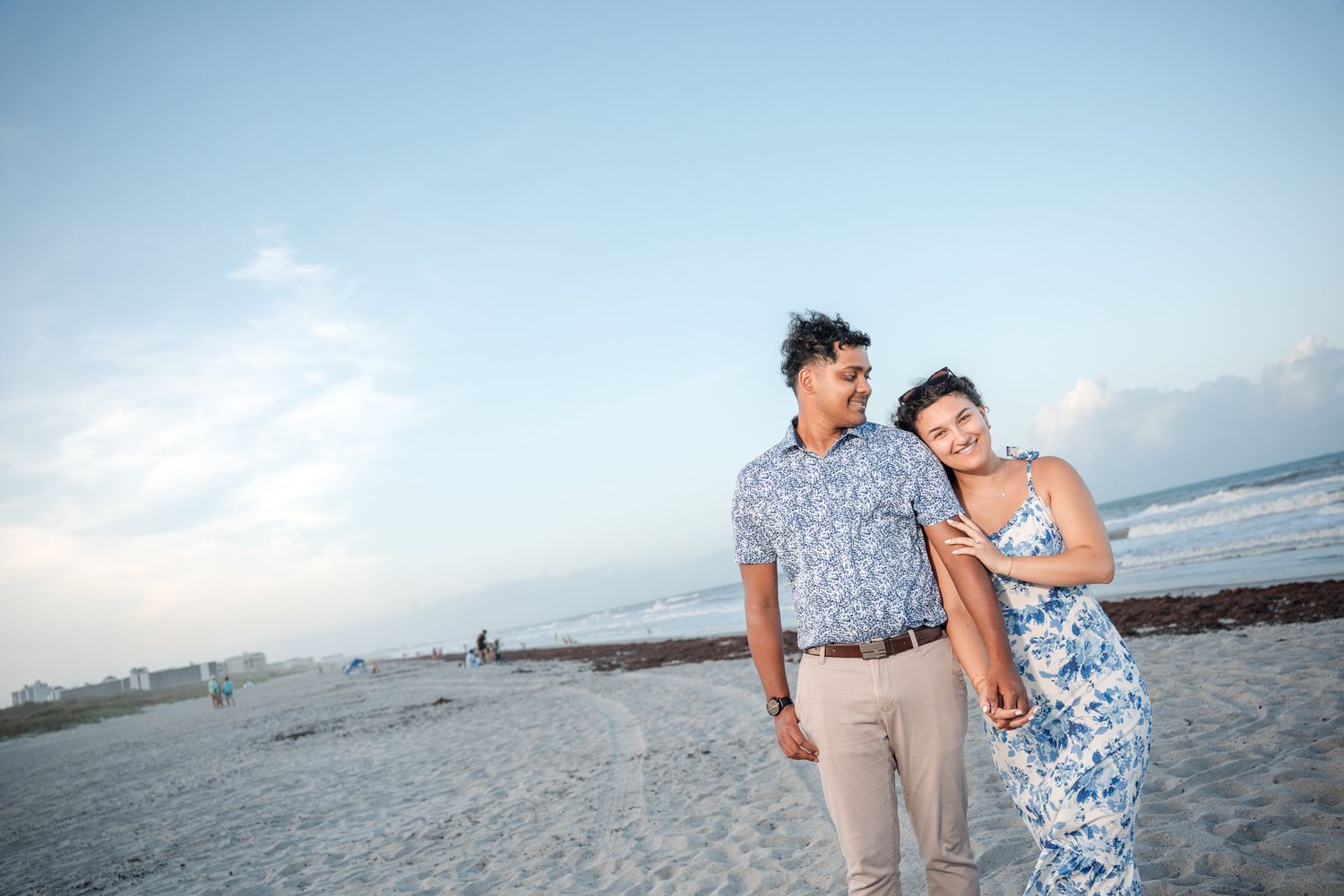 Smiling couple walking on beach at dusk, man in floral shirt, woman in blue floral dress leaning on his shoulder.
