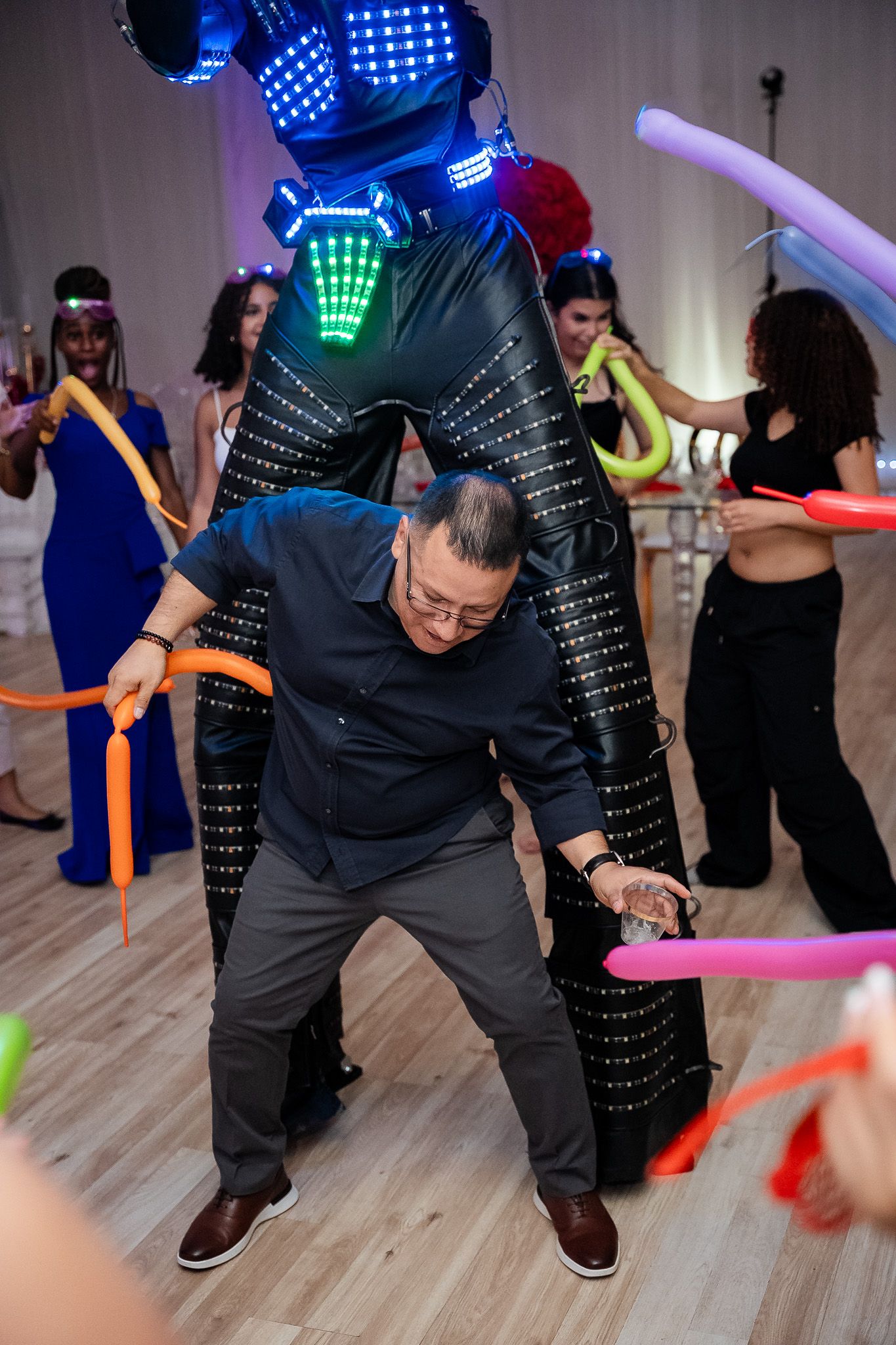 Man doing the limbo under neon balloon sticks at a lively quinceañera party with LED robot.