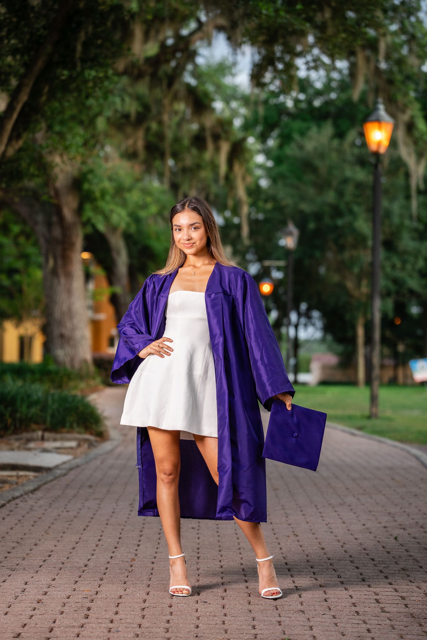 Smiling female graduate in purple gown holds cap while posing on brick pathway lined with trees.