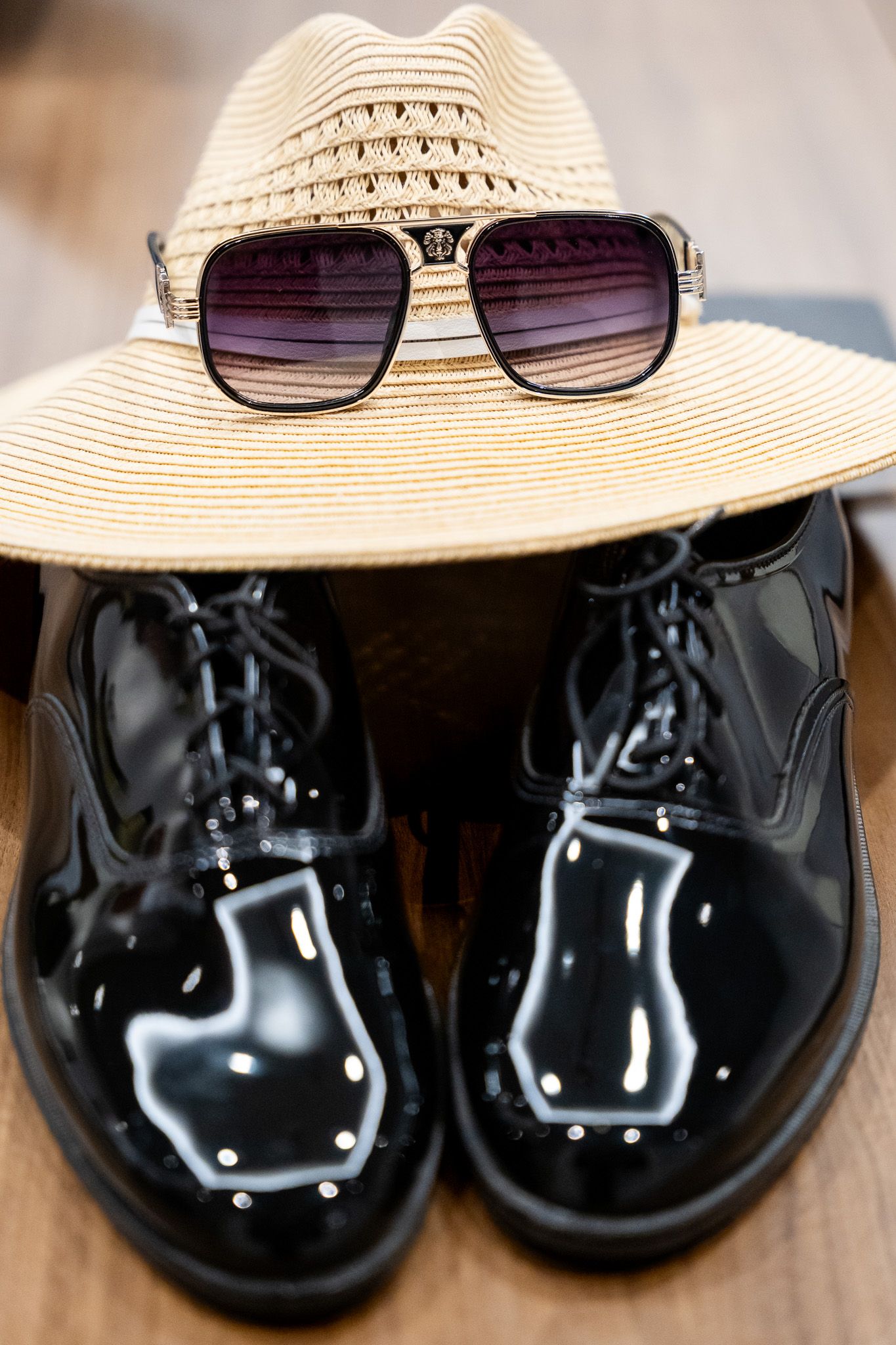 Black patent leather oxford shoes with straw hat and sunglasses on wooden floor.