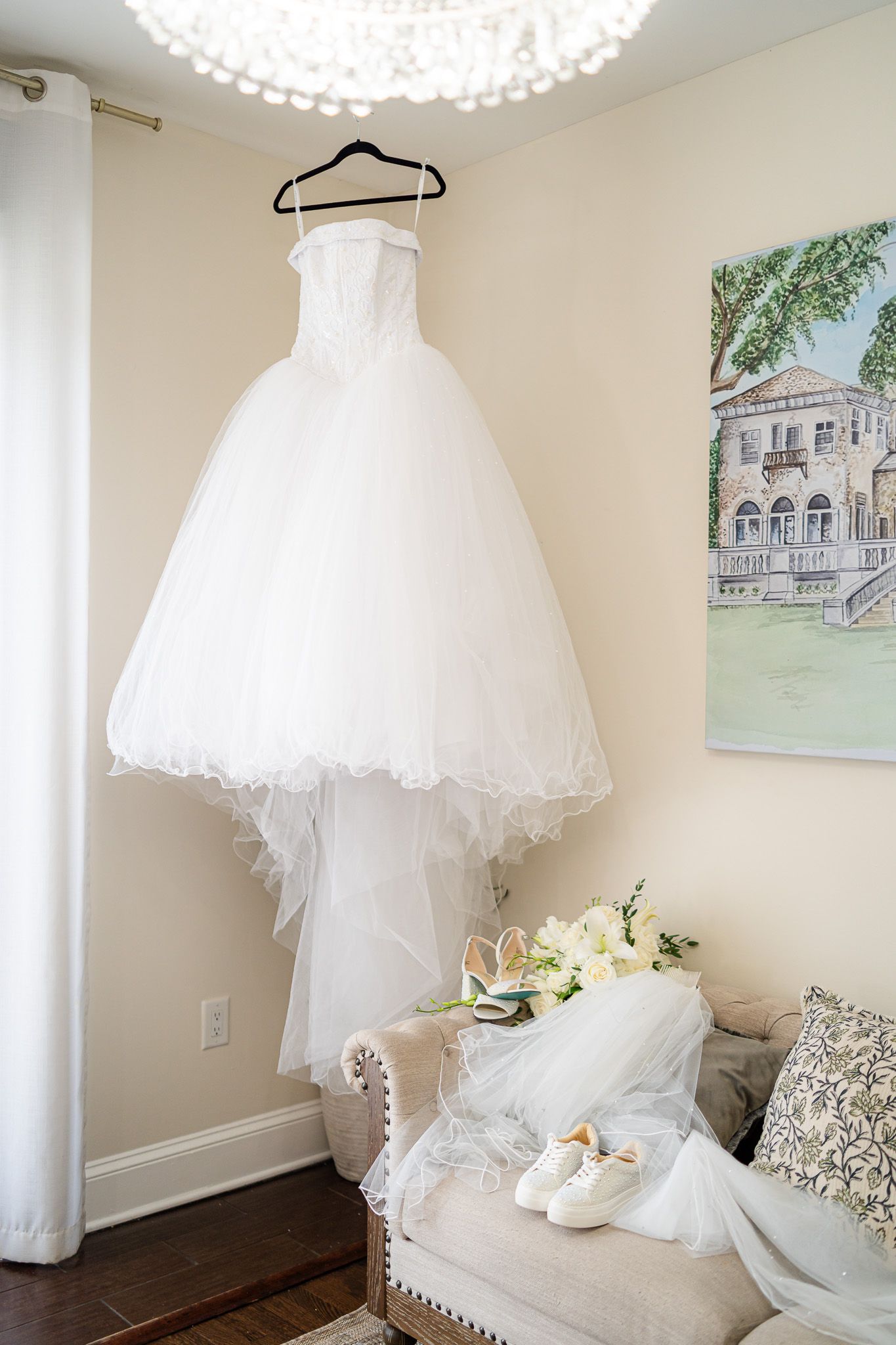Strapless white ballgown wedding dress hanging on hanger with bridal bouquet and sneakers below.