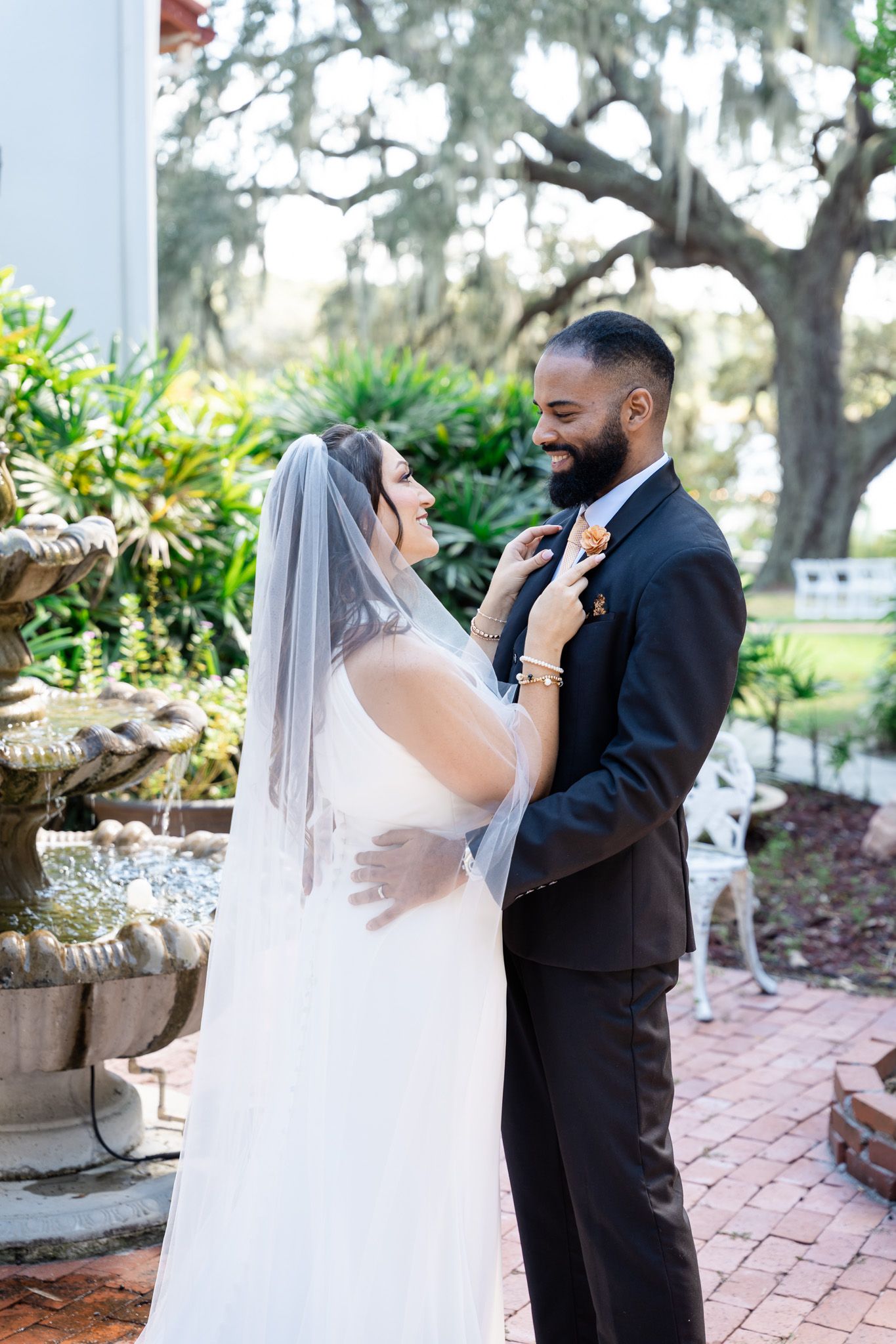 Smiling bride adjusts groom's boutonniere near stone fountain surrounded by lush greenery outdoors.