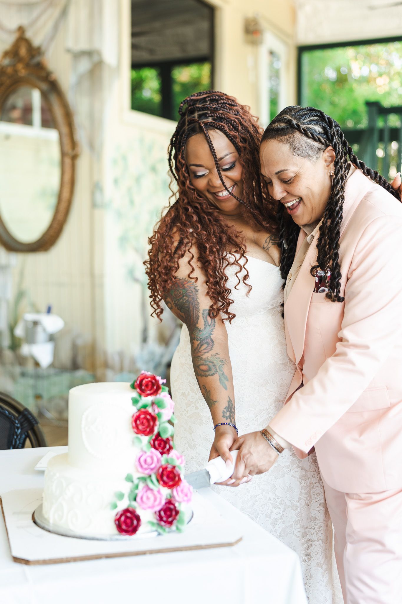 Two brides cutting their white rose-adorned wedding cake, one in a lace dress and one in a pink suit.