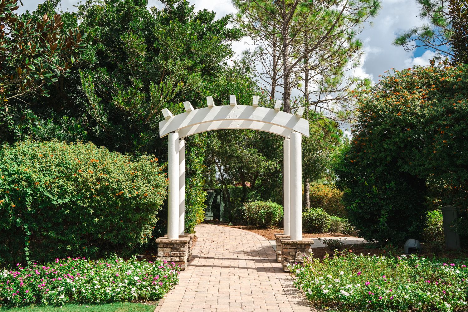 Beautiful white garden pergola archway surrounded by lush greenery and colorful flowers on a brick pathway.