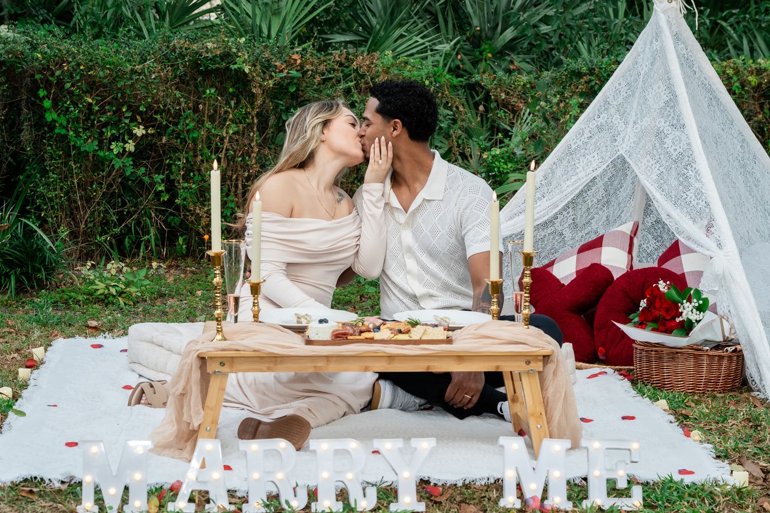 Couple sharing a kiss during romantic outdoor proposal setup with marry me sign and teepee.