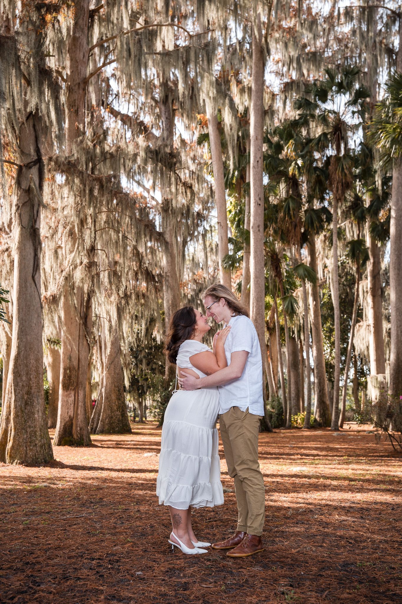 Couple leaning in for a kiss surrounded by tall trees draped with Spanish moss on a sunny day.