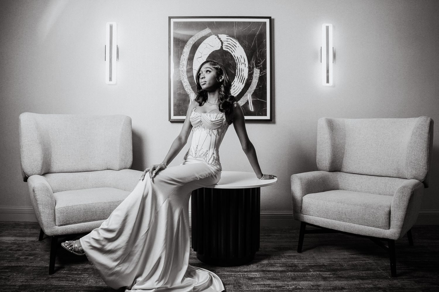 Elegant bride in a flowing white gown poses gracefully on a dark table between two chairs in a stylish room.