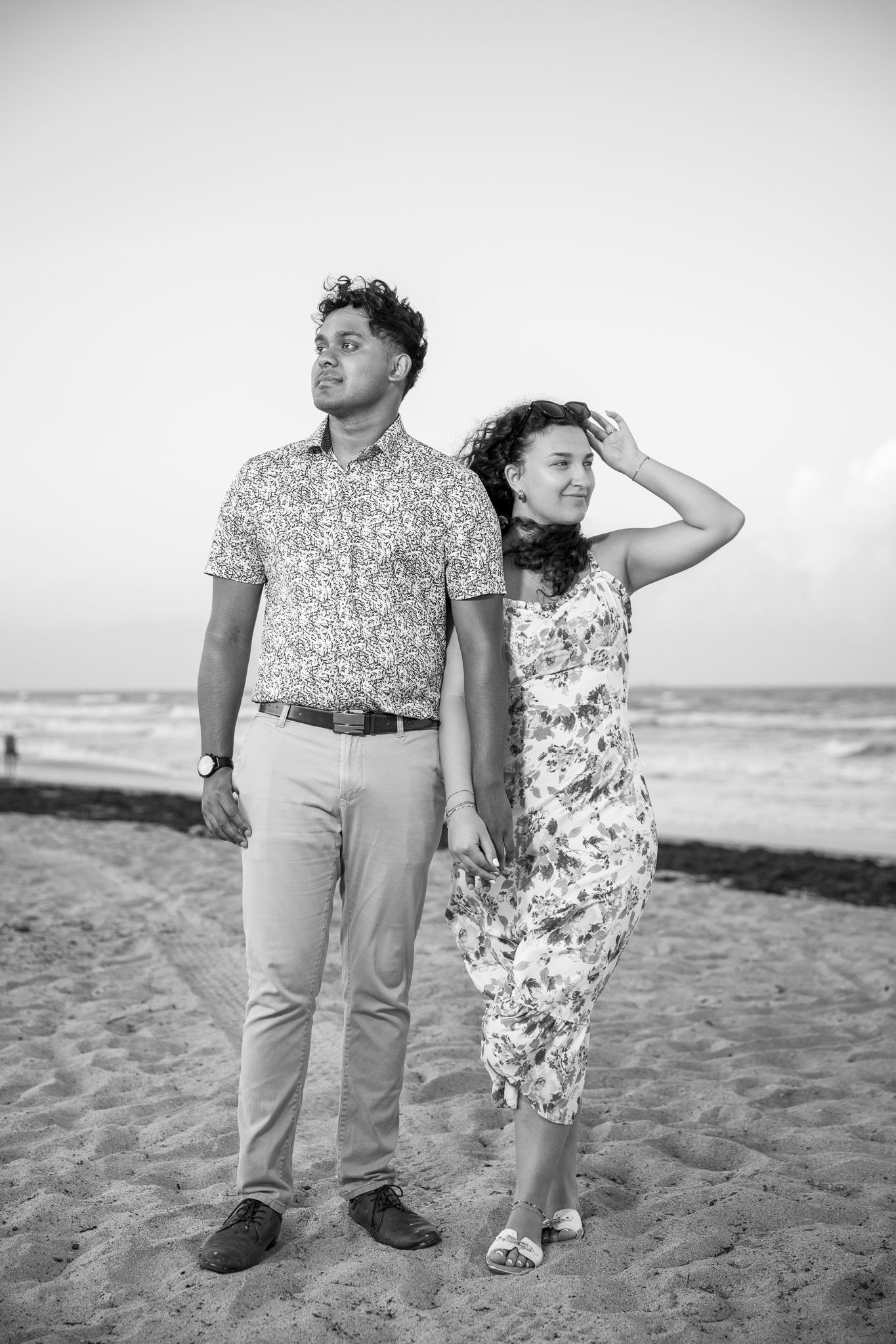 Black and white photo of a couple holding hands on a beach, woman touching her hair in the breeze.