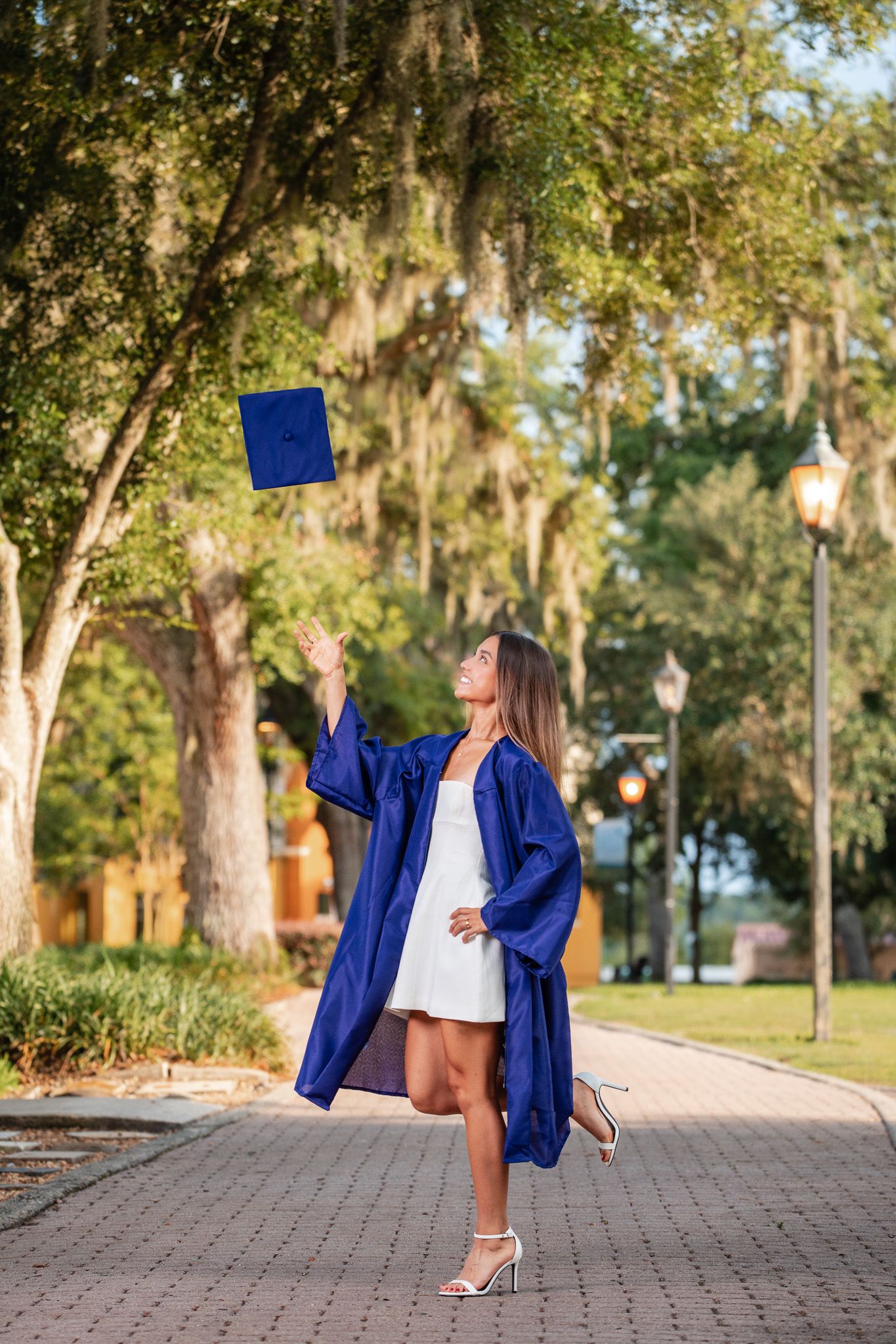 Graduate in blue cap and gown tosses mortarboard into the air on a scenic campus walkway.