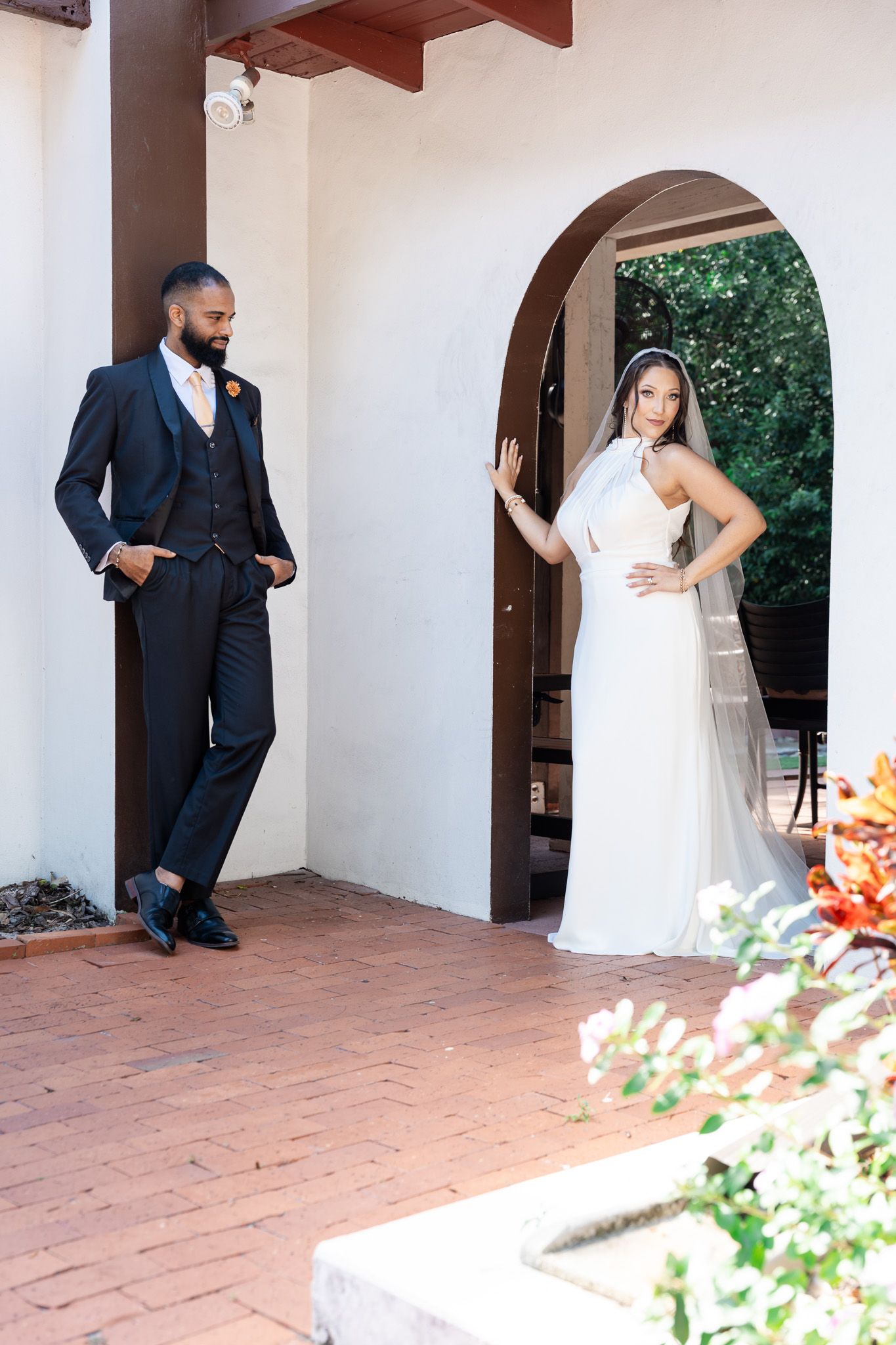 Groom in navy suit leans against white arch wall as bride in halter gown poses in doorway.