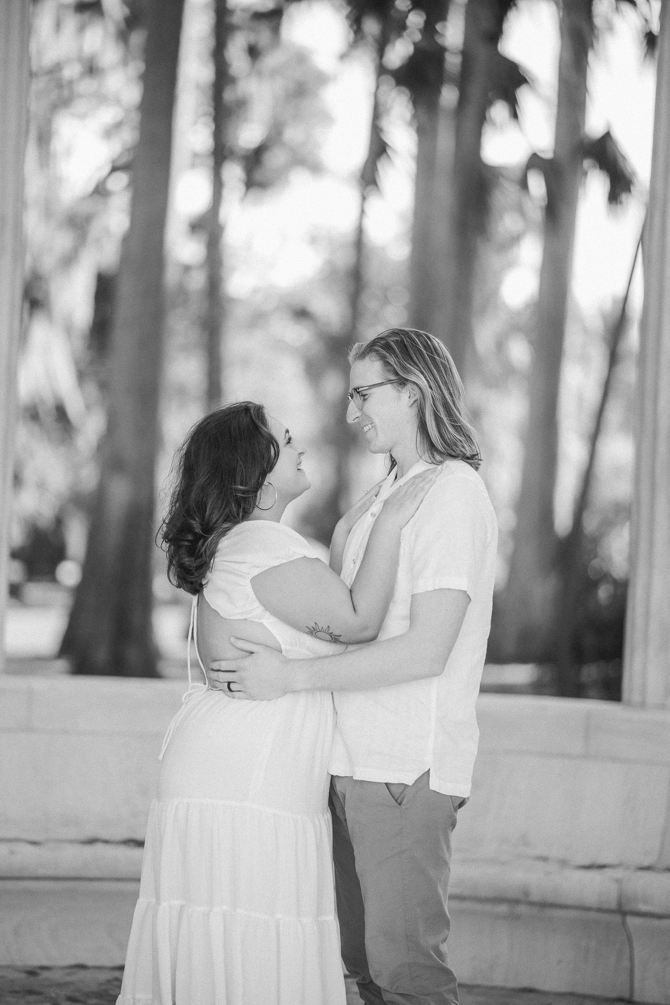 Black and white photo of a laughing couple embracing near white columns with palm trees in background.