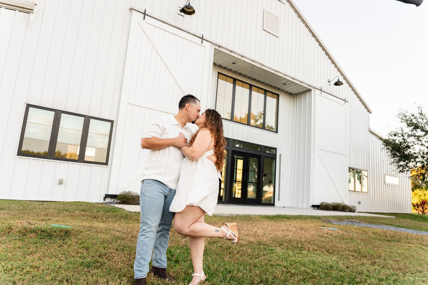 Couple dancing and laughing on green grass in front of a white barn at sunset.