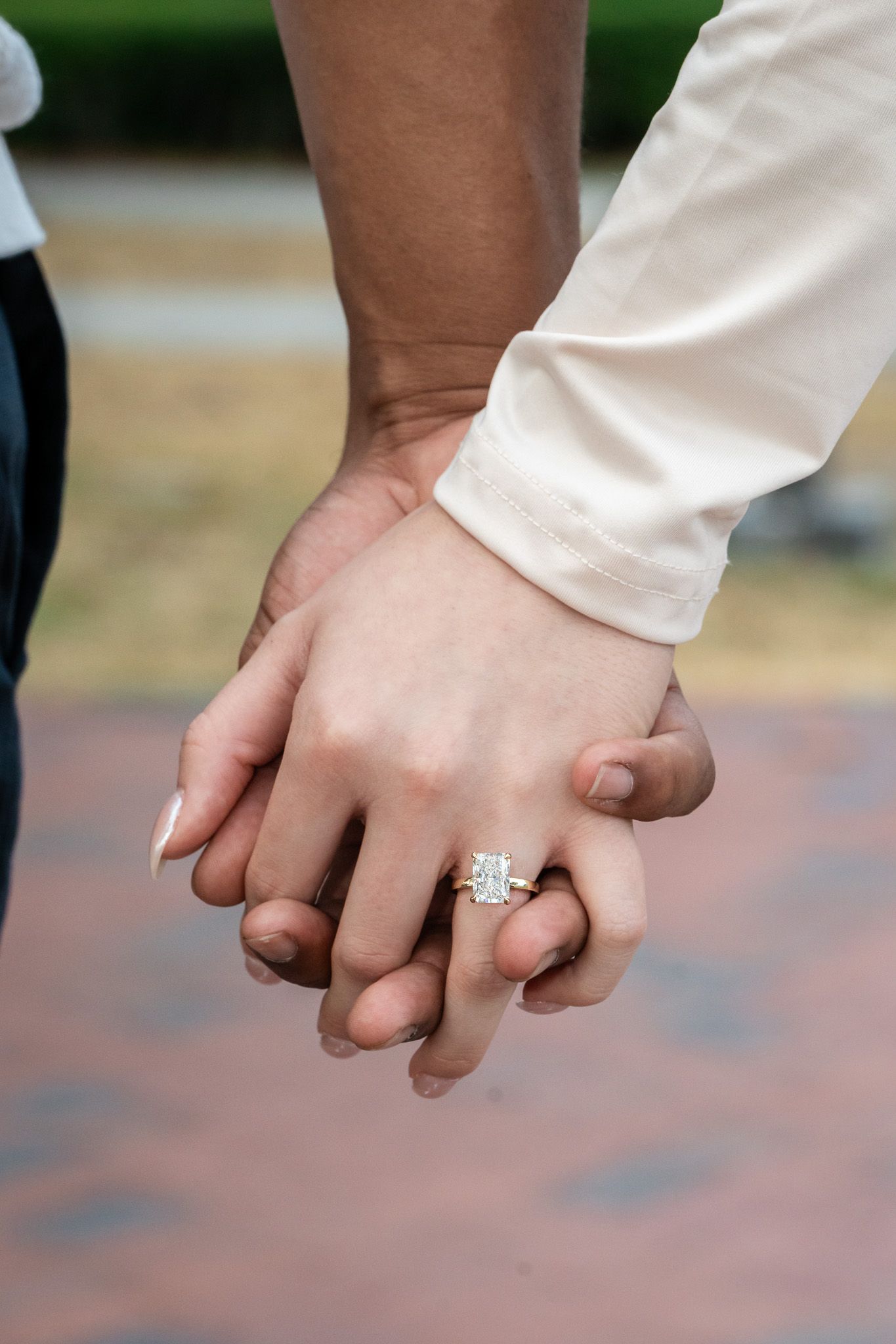 Close-up of couple holding hands displaying a diamond engagement ring on brick walkway.