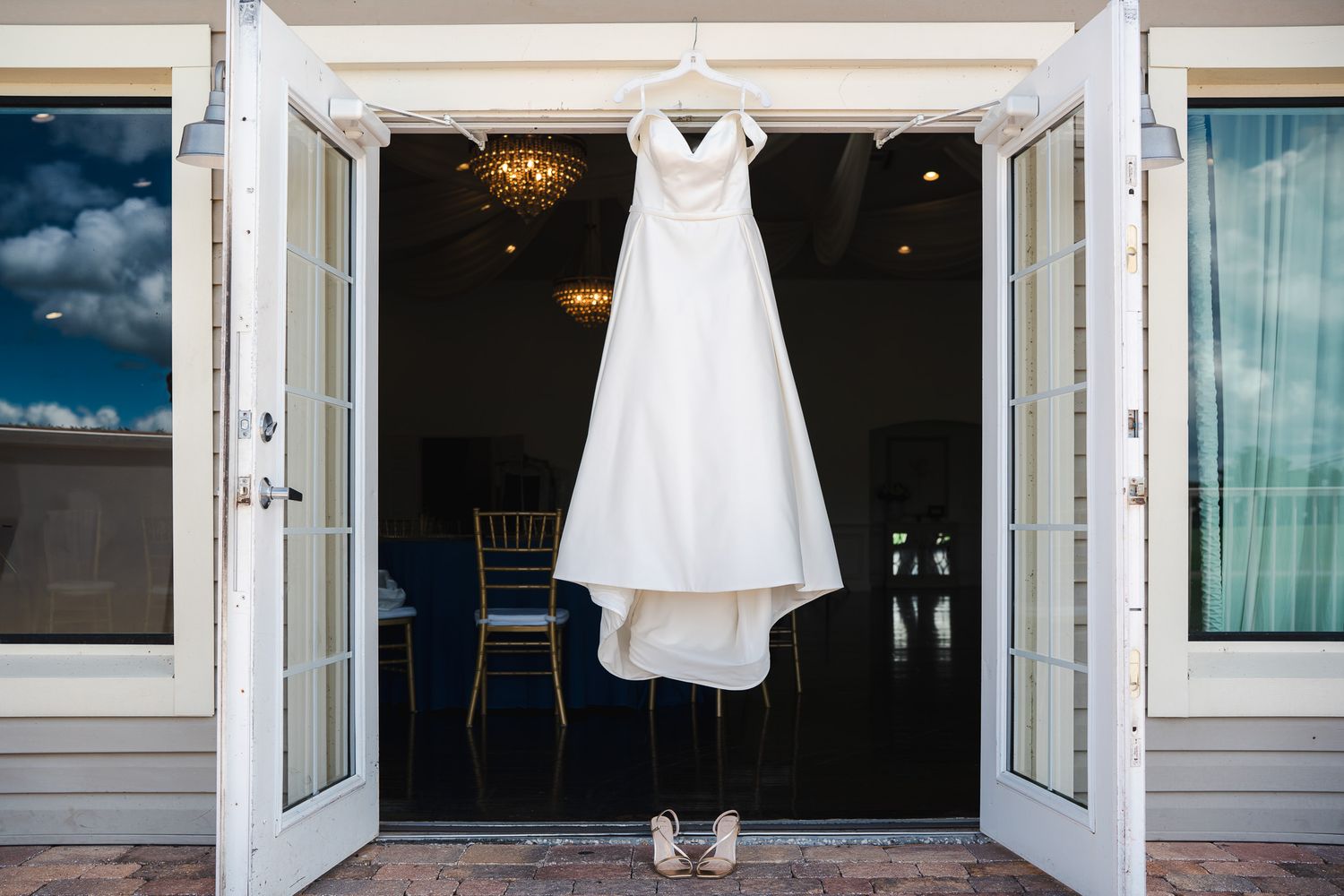 Elegant white wedding gown hanging on French doors with bridal heels below in a decorated venue.