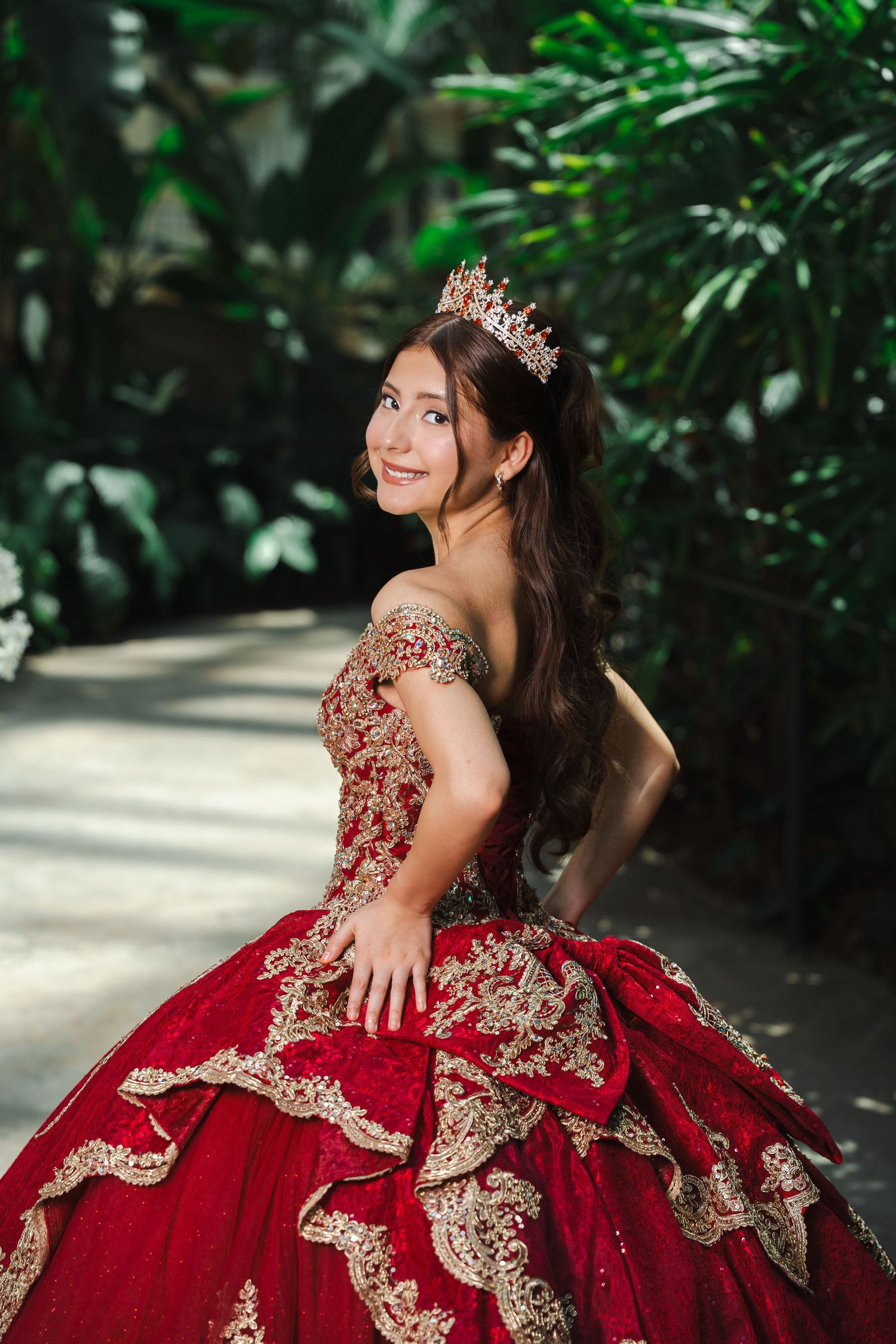 Young woman in red gold quinceañera gown and crown poses gracefully in lush garden setting.