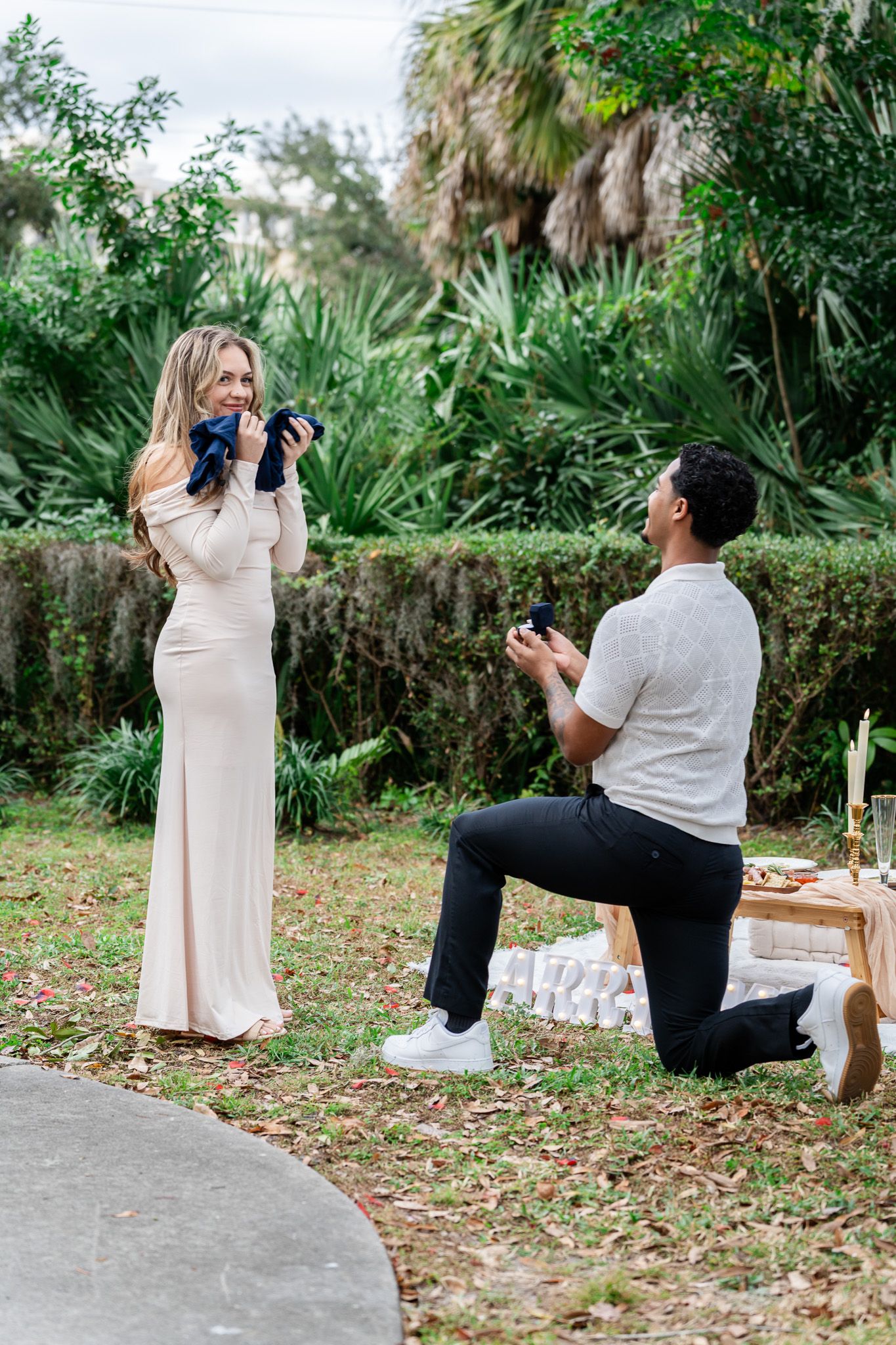 Man proposes on one knee to surprised woman in blush gown at outdoor garden setting.