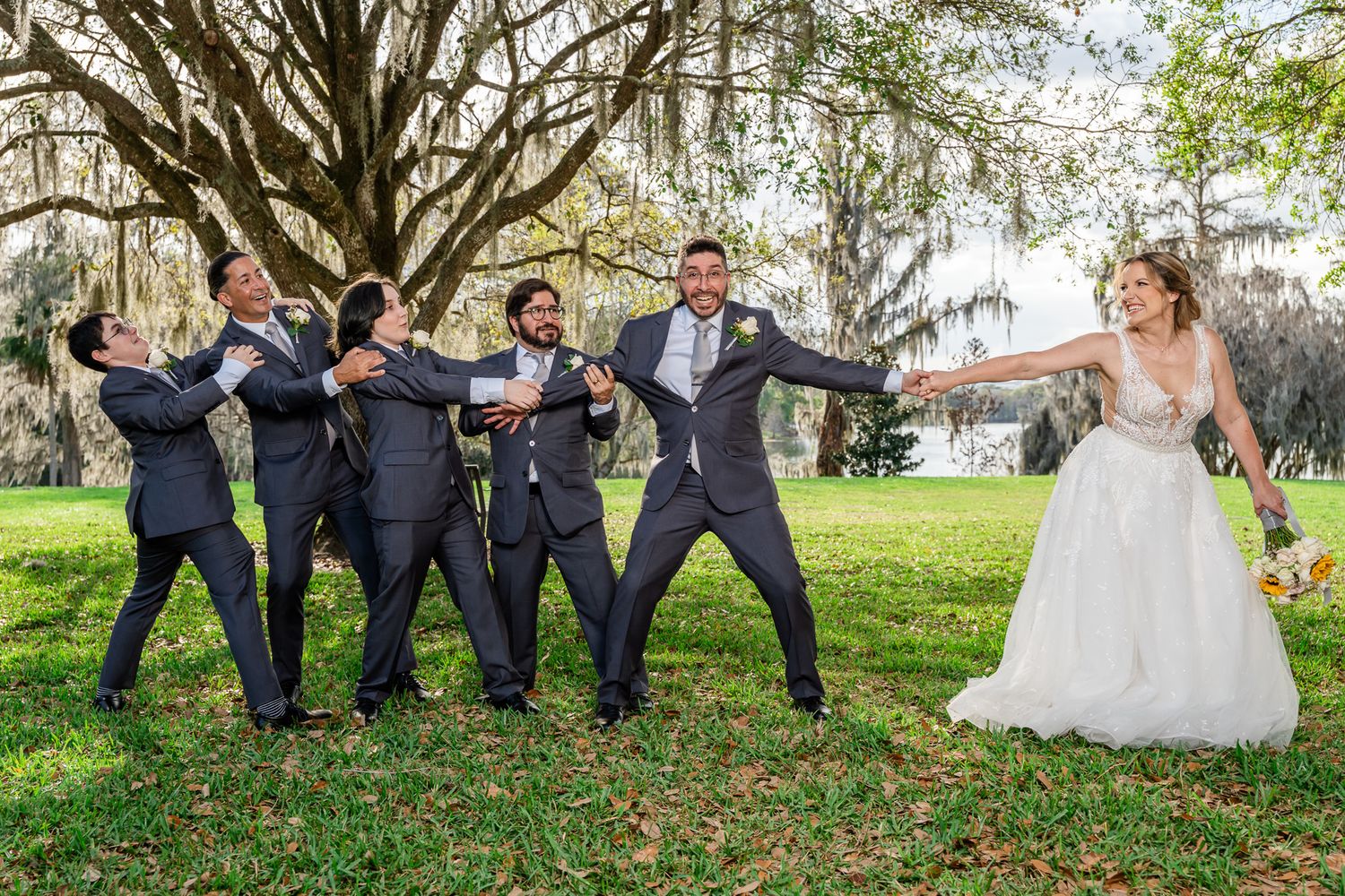 Bride pulls groom away as groomsmen in gray suits dramatically reach out in playful outdoor wedding party photo.