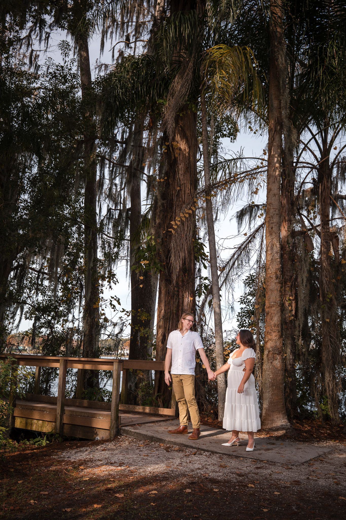 Engaged couple holding hands on a wooden walkway surrounded by tall moss-covered trees.