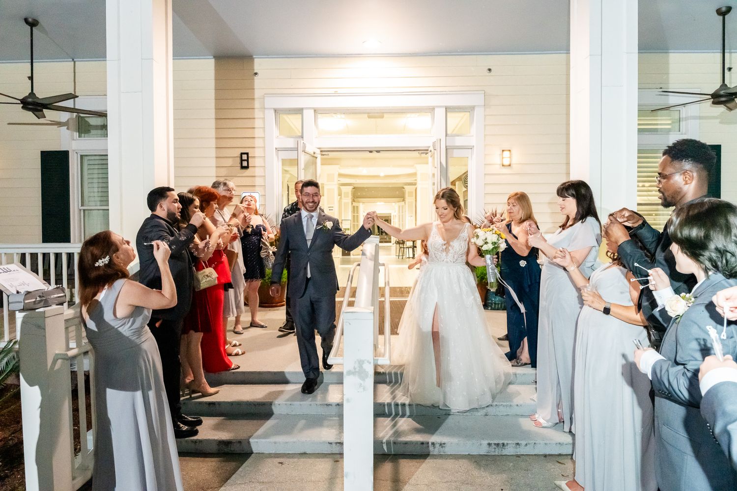 Bride and groom make a grand exit through cheering wedding guests on a decorated venue porch.