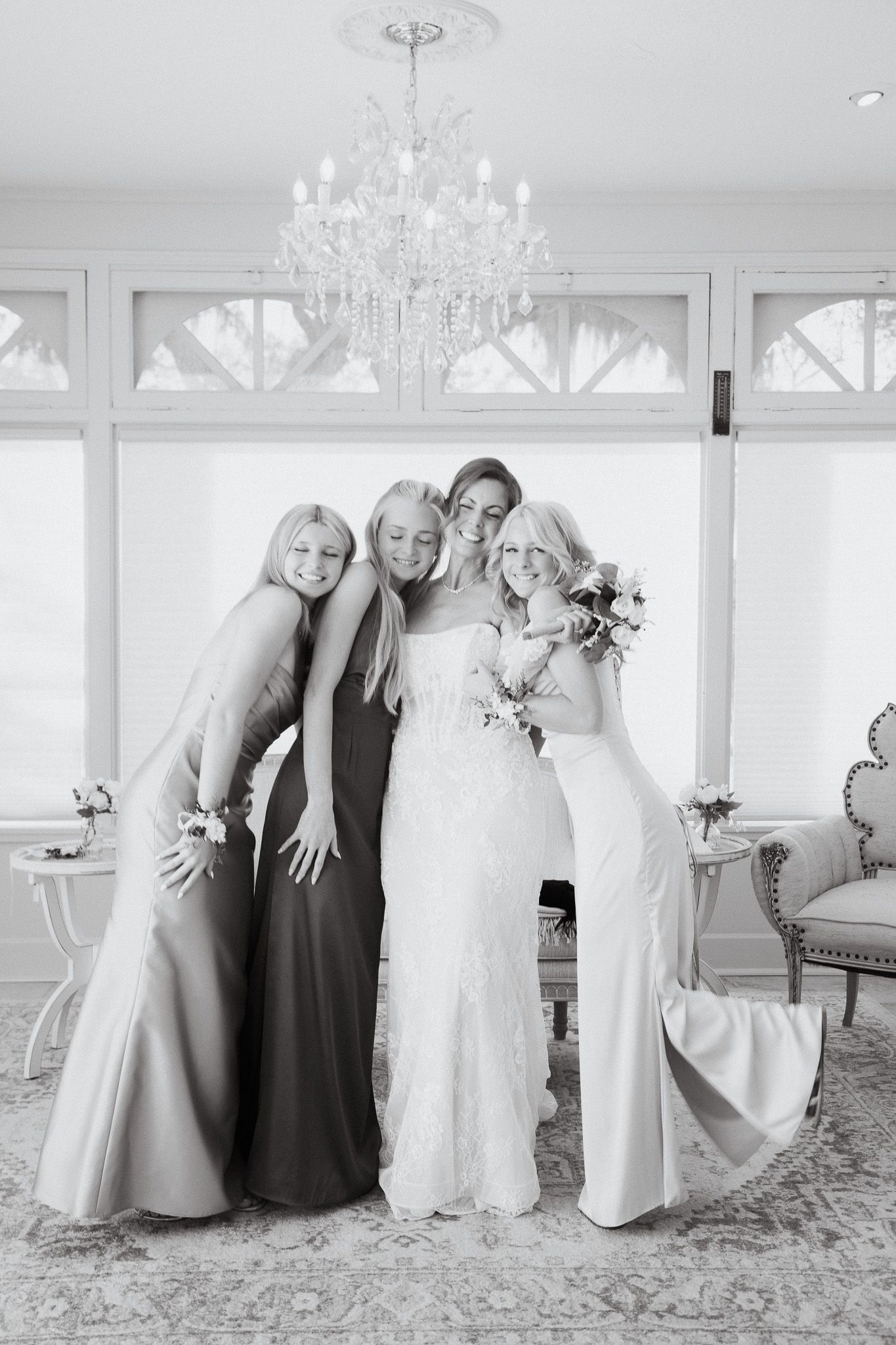 Bride poses with three bridesmaids in elegant gowns under a crystal chandelier in a bright white room.
