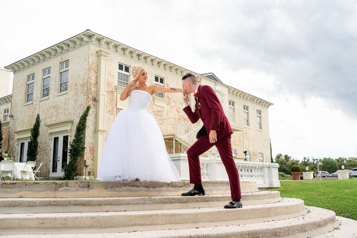 Bride in white gown and groom in burgundy suit pose playfully on mansion steps.