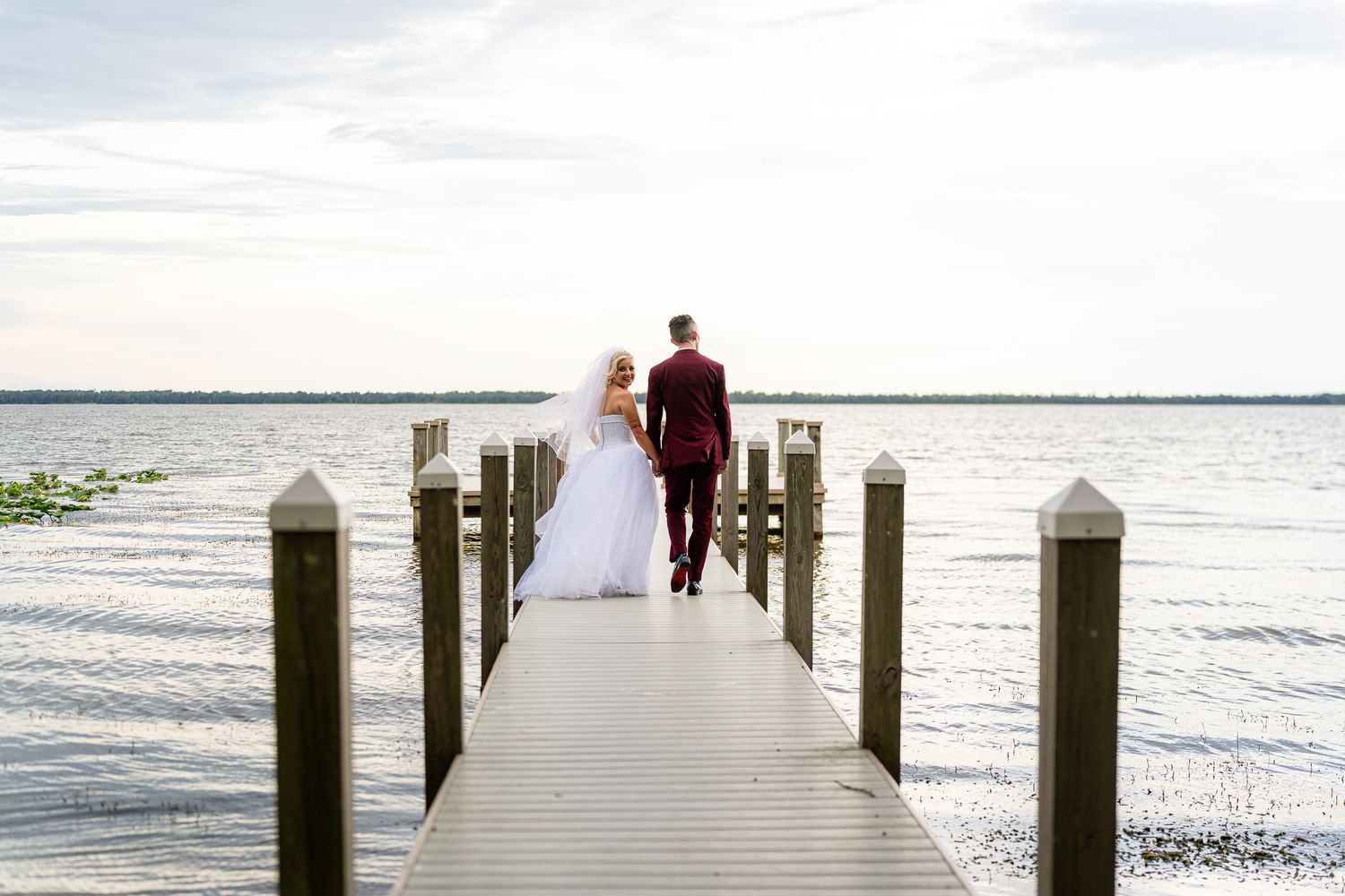 Newlyweds walking hand in hand down wooden pier toward serene open water.