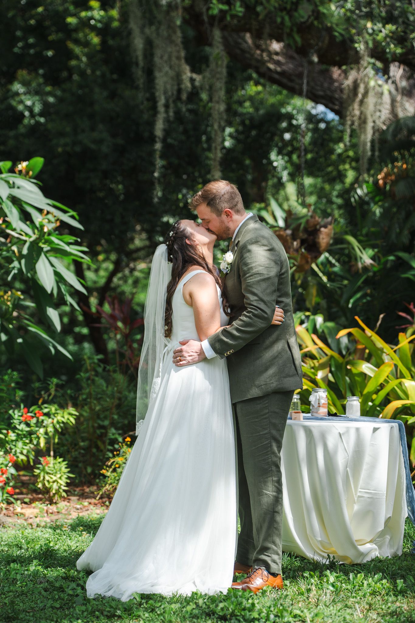 Bride and groom share their first kiss during an outdoor garden wedding ceremony with lush green foliage backdrop.