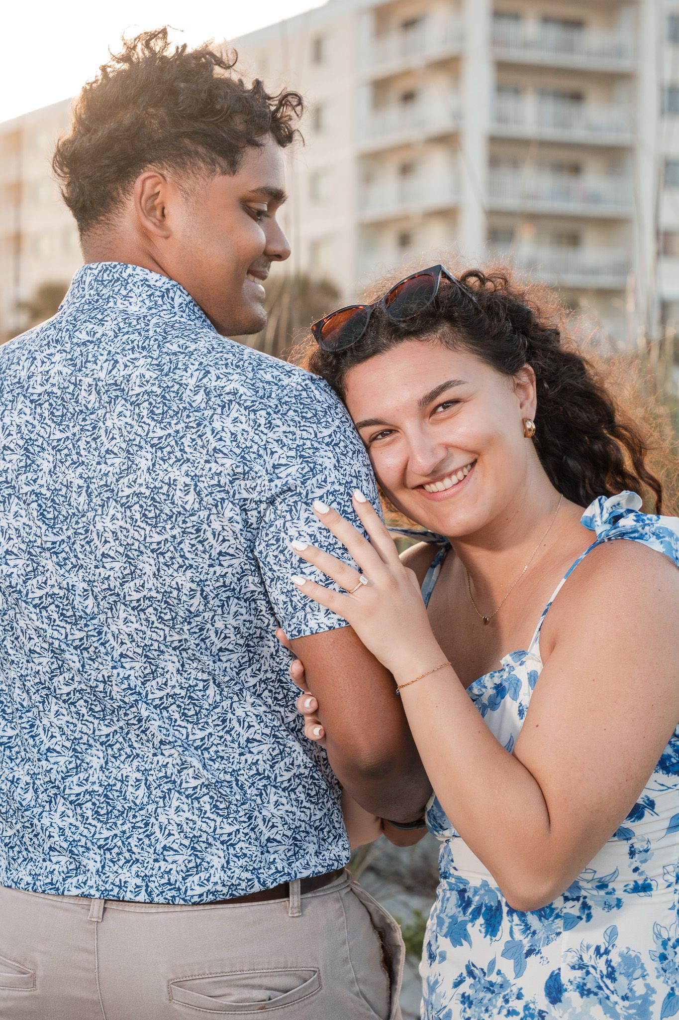 Smiling woman in floral dress hugs man in blue patterned shirt outdoors near beach building at sunset.