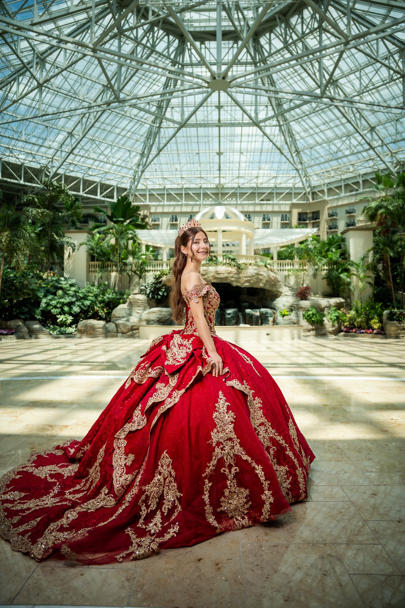 Young woman in stunning red and gold embroidered ballgown quinceañera dress poses in elegant glass atrium with tropical plants.
