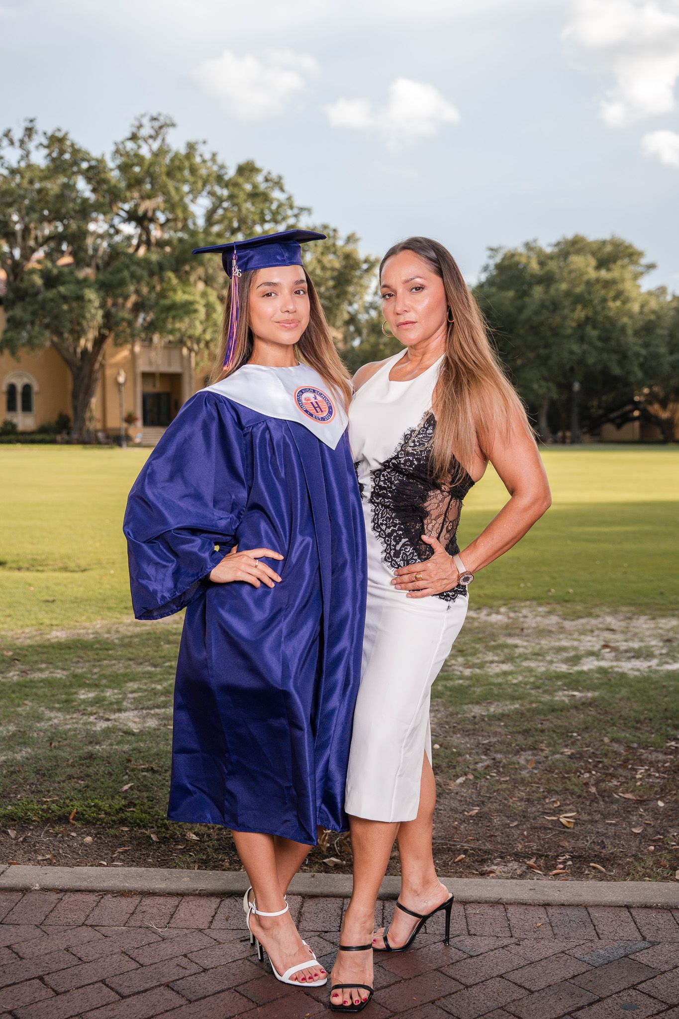 Young female graduate in blue cap and gown stands proudly with her mother in white dress outdoors.
