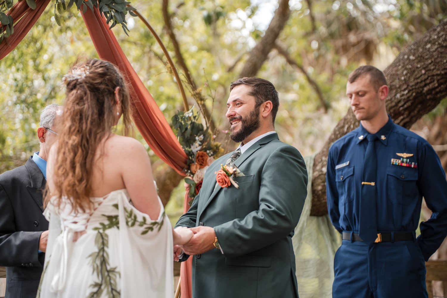 Groom smiling at bride during outdoor wedding ceremony with groomsman in Coast Guard uniform standing nearby.
