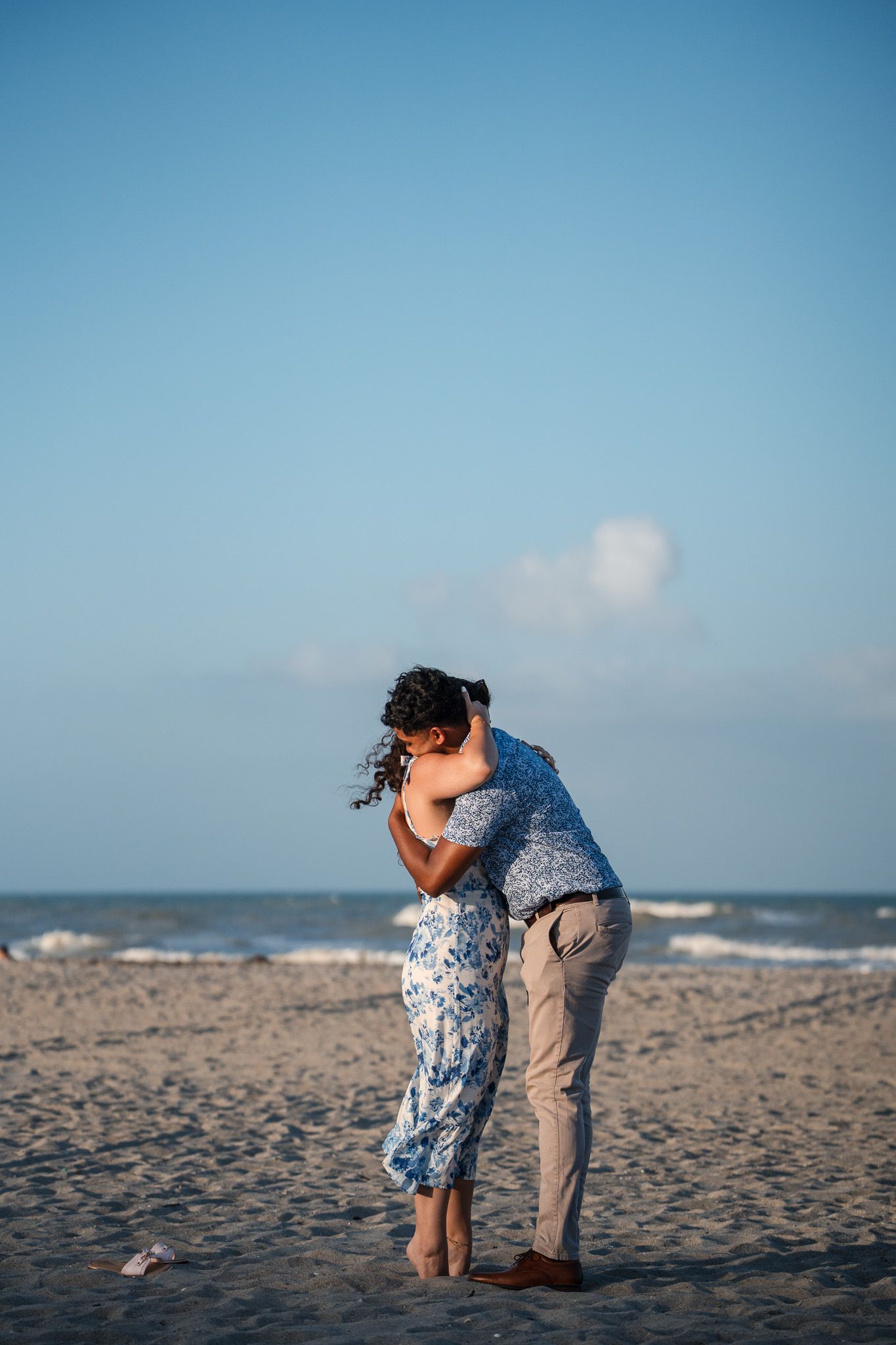 Romantic couple embracing and kissing on a sandy beach with calm ocean waves and blue sky backdrop.