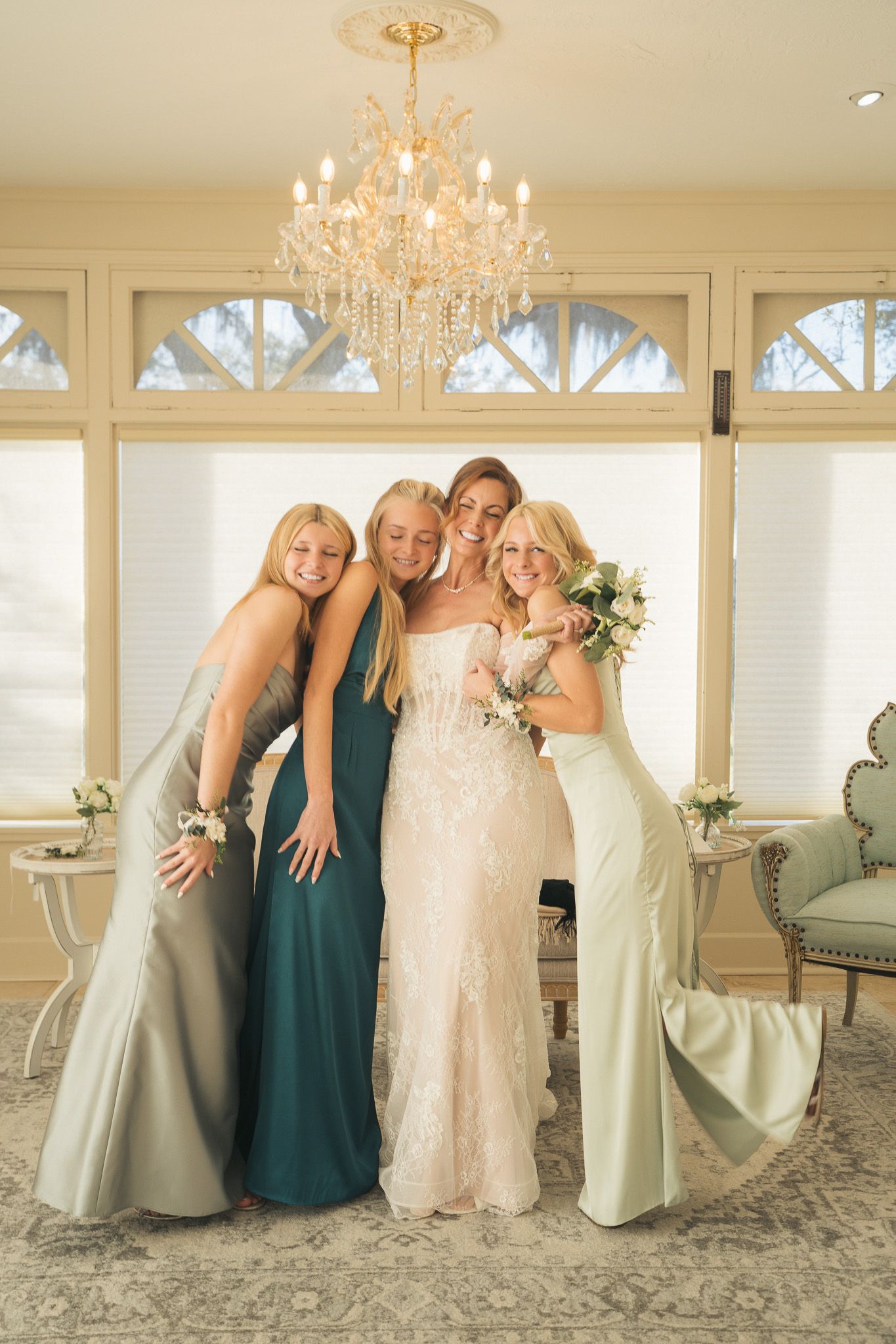 Bride poses with three bridesmaids in sage and teal gowns under elegant chandelier.