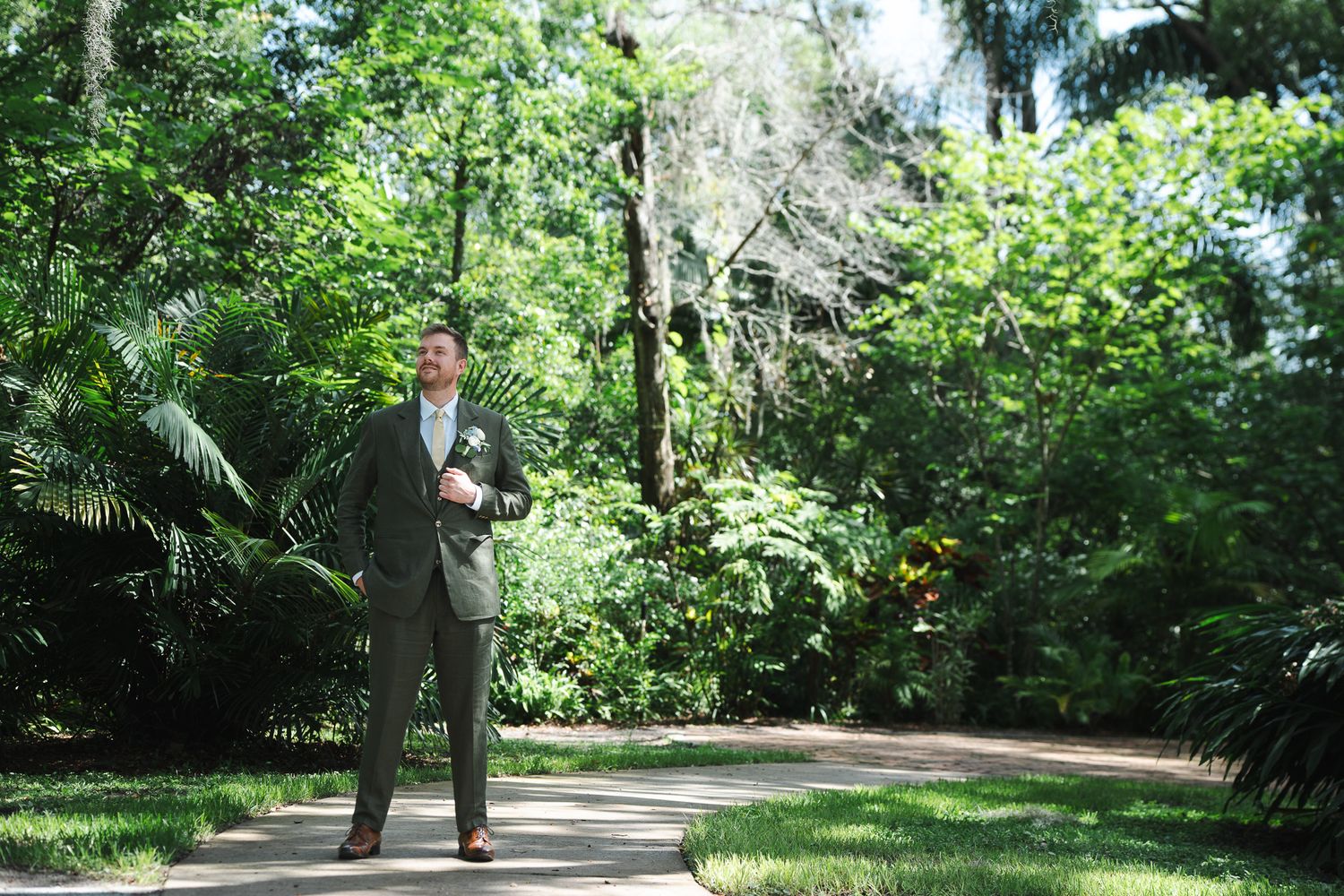 Handsome groom in olive suit standing on garden pathway surrounded by lush greenery.