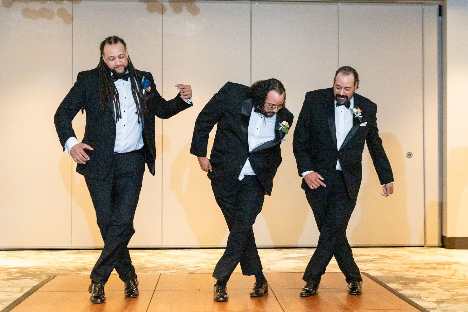 Three groomsmen in black tuxedos strike fun dance poses on ballroom dance floor.