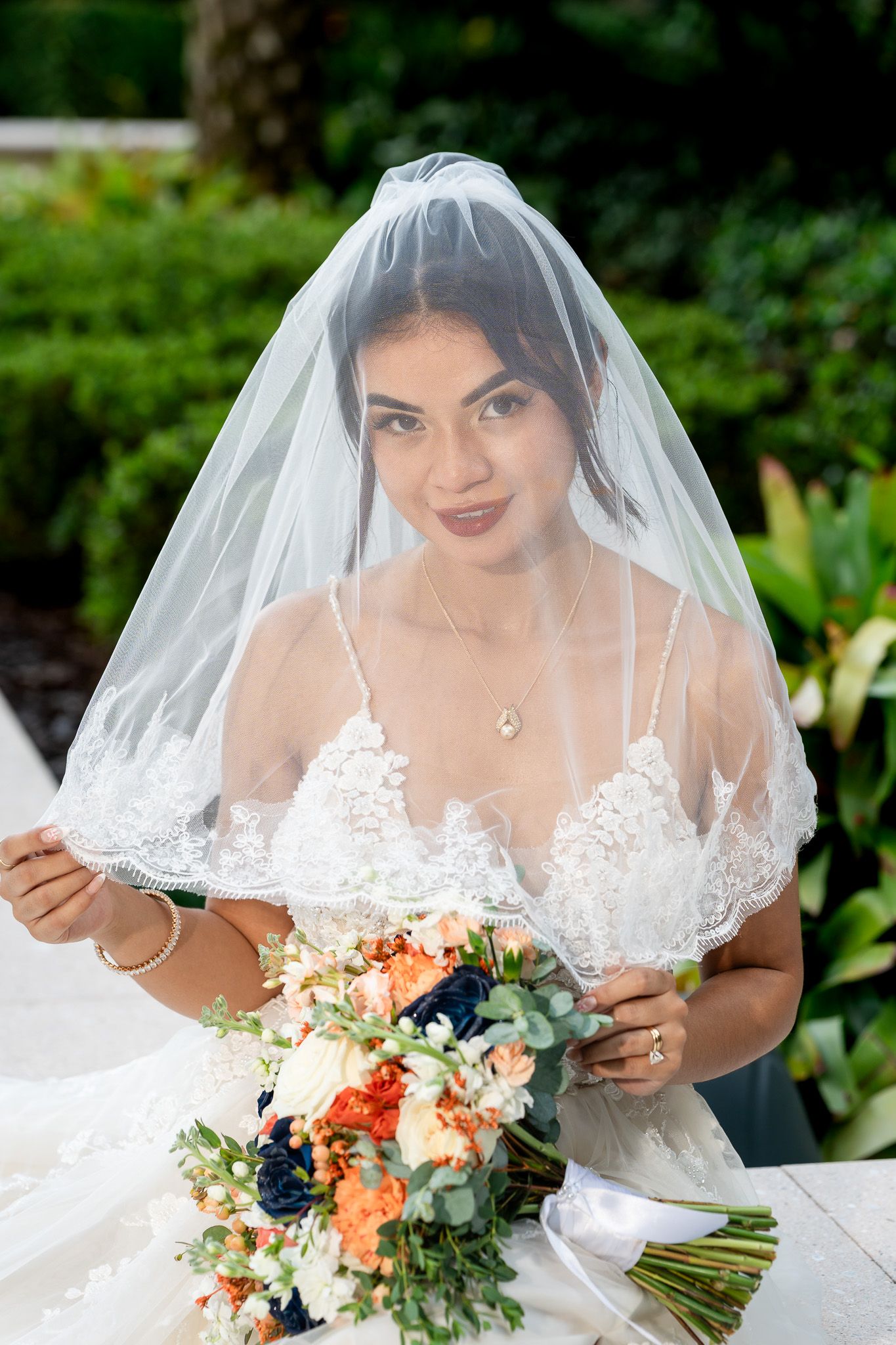 Beautiful bride in lace gown holding colorful bouquet with veil over face outdoors.