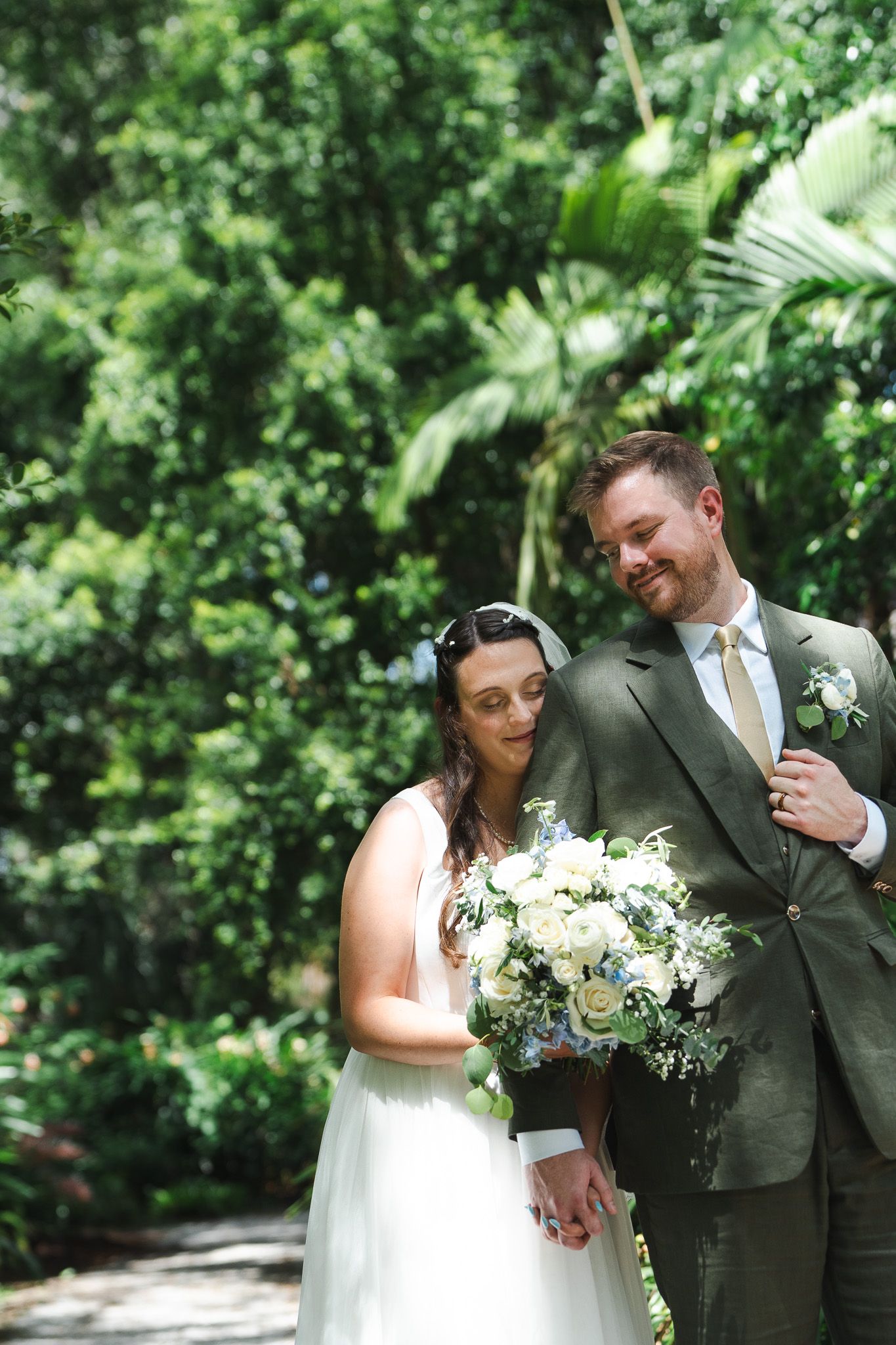 Bride and groom hold hands while posing with a white floral bouquet in a lush garden setting.