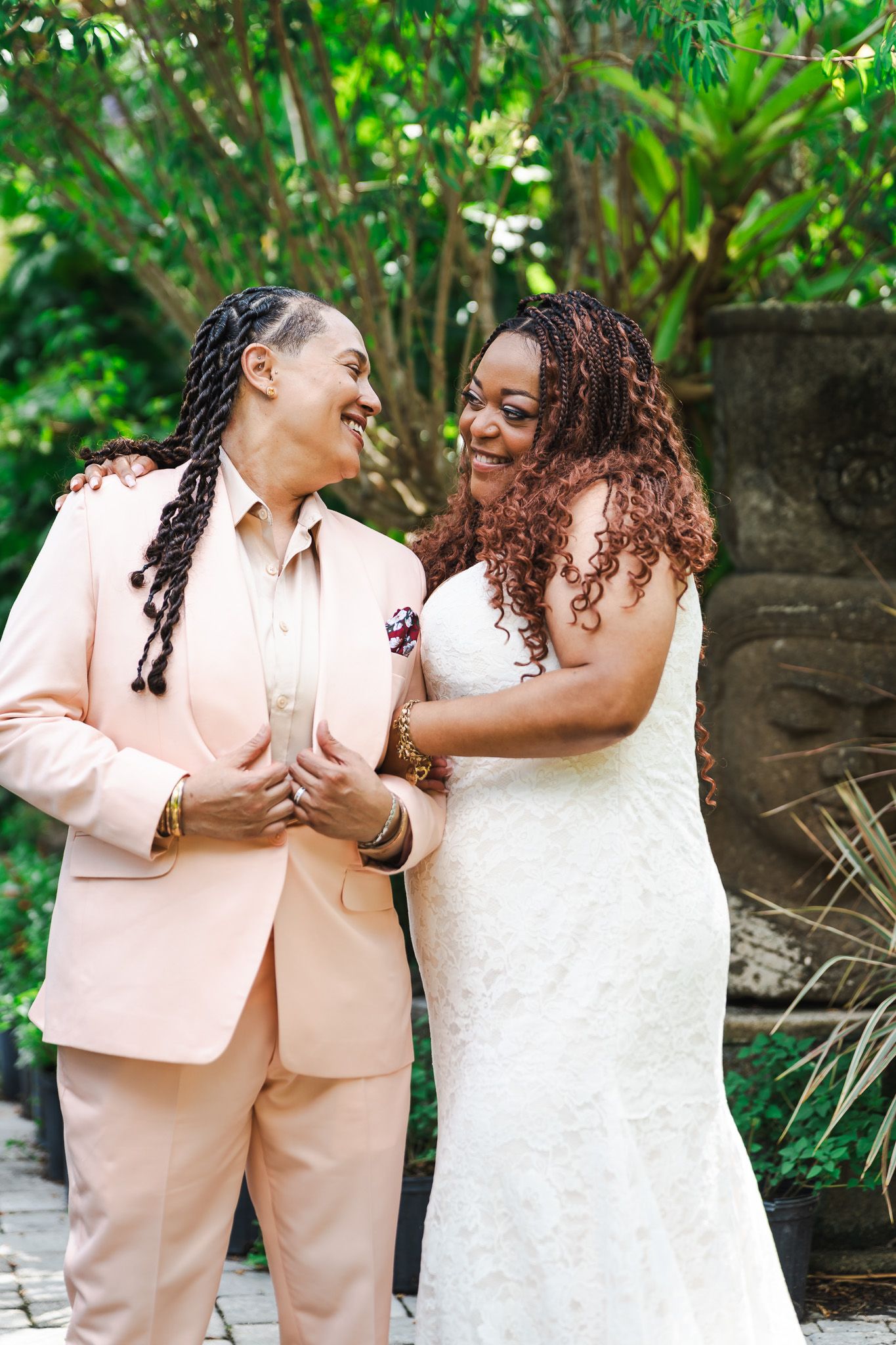 Two brides laugh together in garden wearing pink suit and white lace wedding gown.