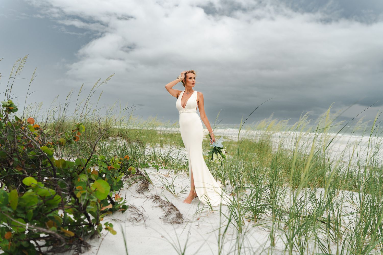 Elegant bride in white fitted gown with front slit holding bouquet while standing on sandy beach dunes.