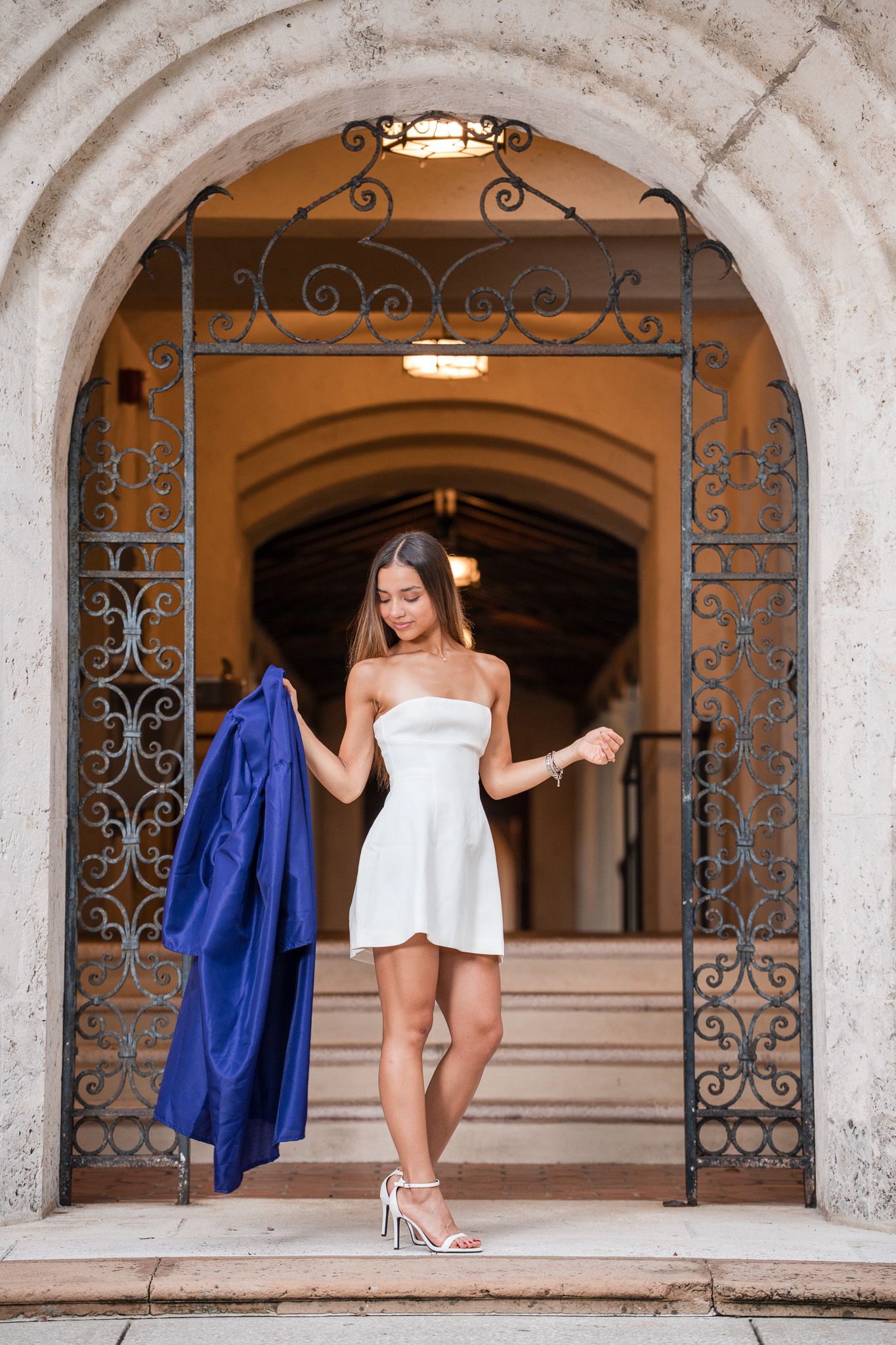 Young woman in white strapless dress holding blue graduation gown under ornate iron arch doorway.