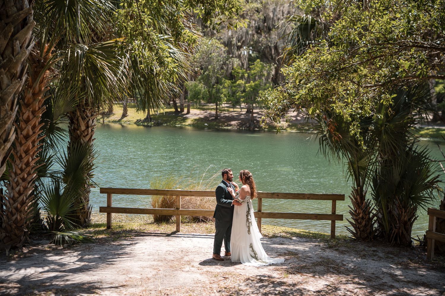 Bride and groom share a romantic moment by a serene lake surrounded by lush trees at an outdoor wedding venue.