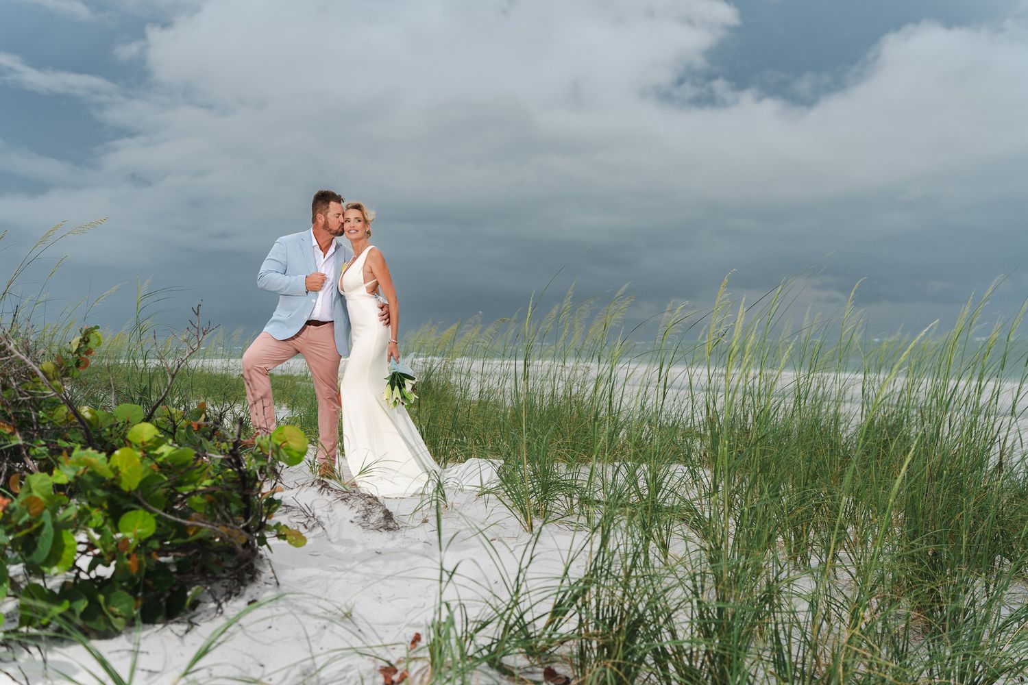 Newlyweds embrace on sandy beach dunes with stormy skies and sea grass in background.