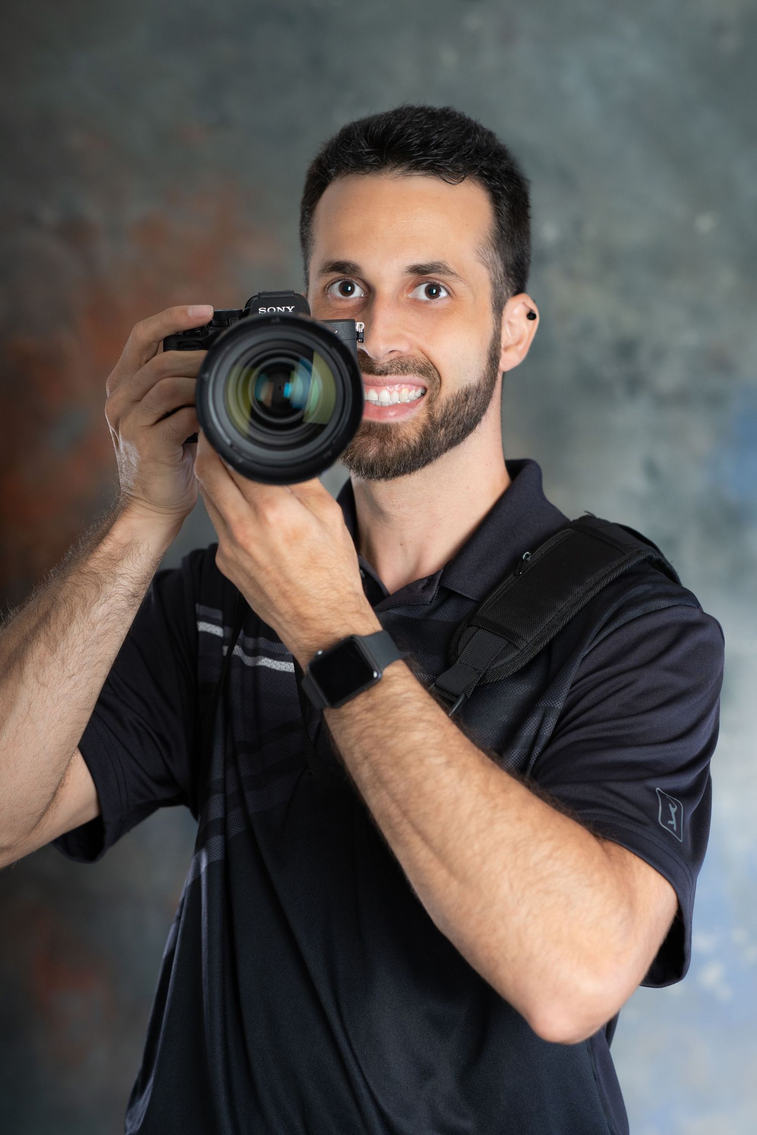 Smiling male photographer holding a Sony camera ready to shoot against a textured gray studio background.