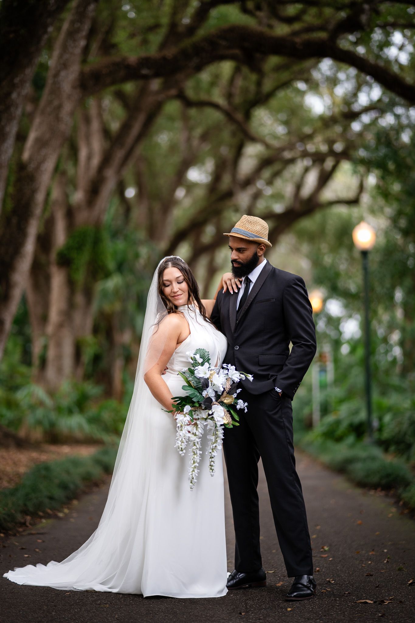 Elegant bride and groom stand together under a canopy of trees, bride holding a cascading white bouquet.