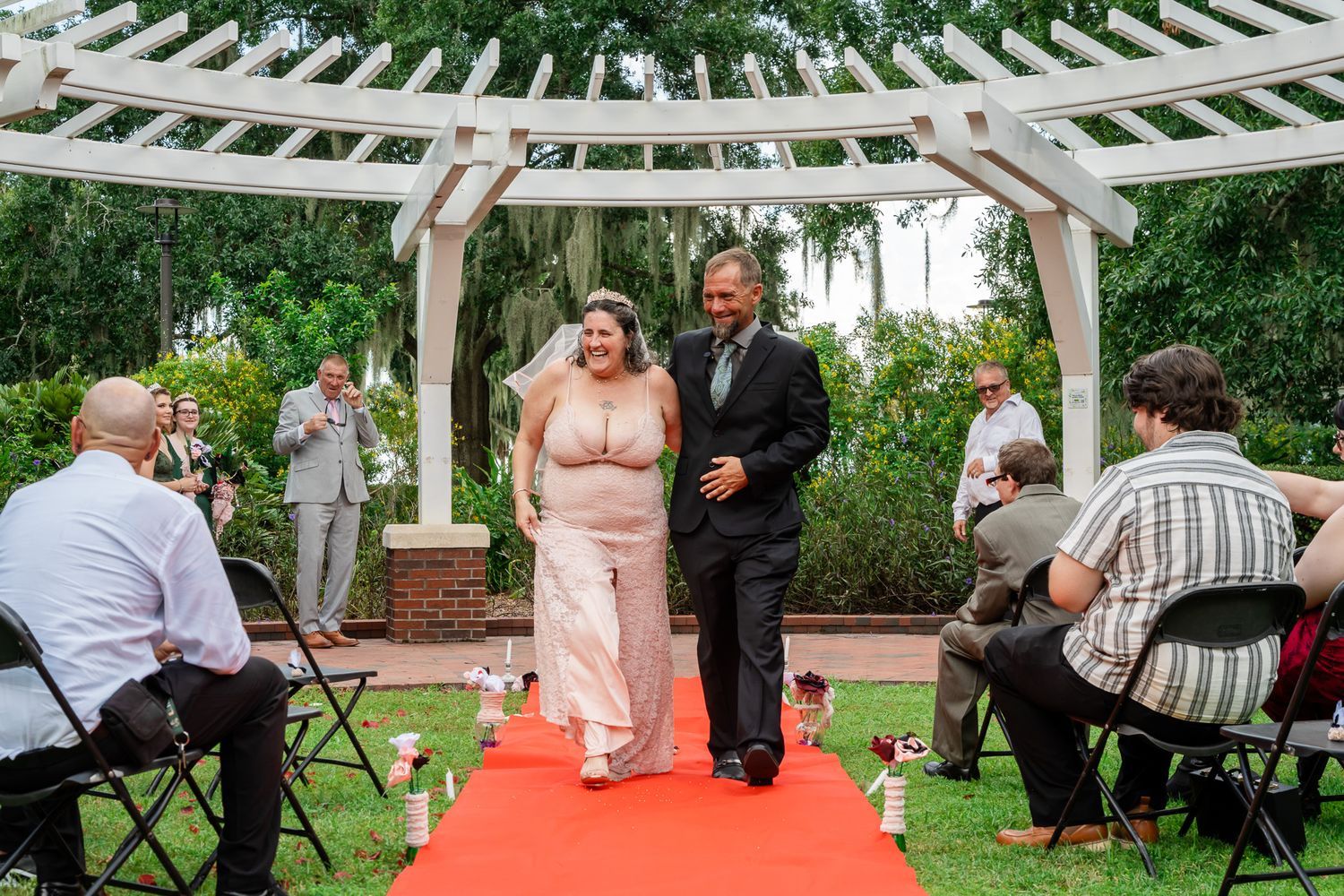 Newlywed couple walks down red carpet aisle under white pergola at outdoor garden wedding ceremony.