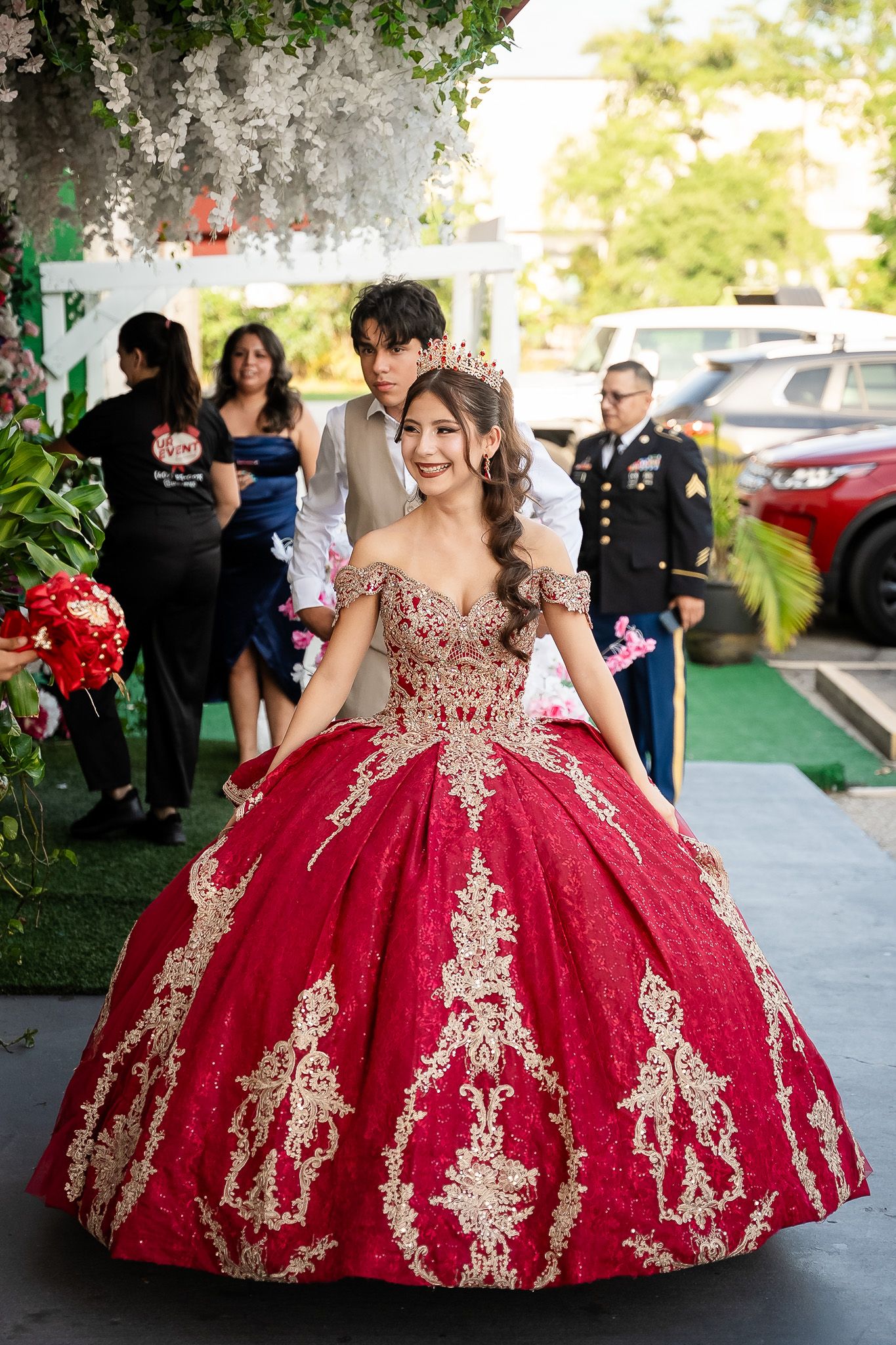 Young woman in red and gold quinceañera ball gown with crown walks at outdoor celebration.