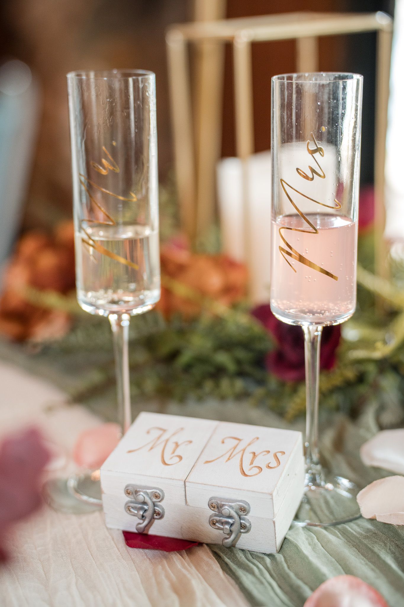 Mr. and Mrs. champagne flutes with gold script beside a white wedding ring box on a decorated table.
