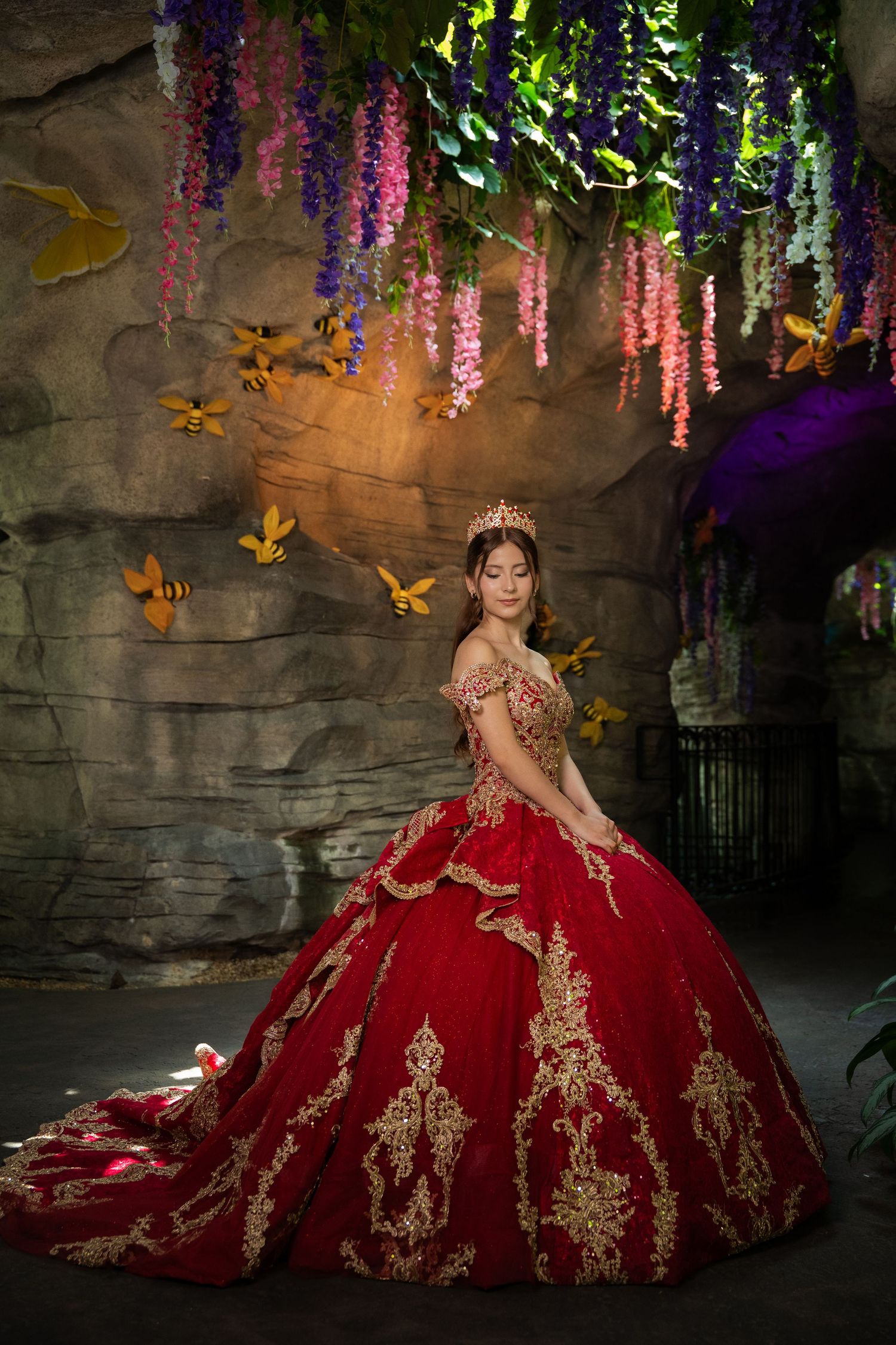 Young woman in red and gold quinceañera ball gown with crown poses among hanging wisteria and golden butterflies.