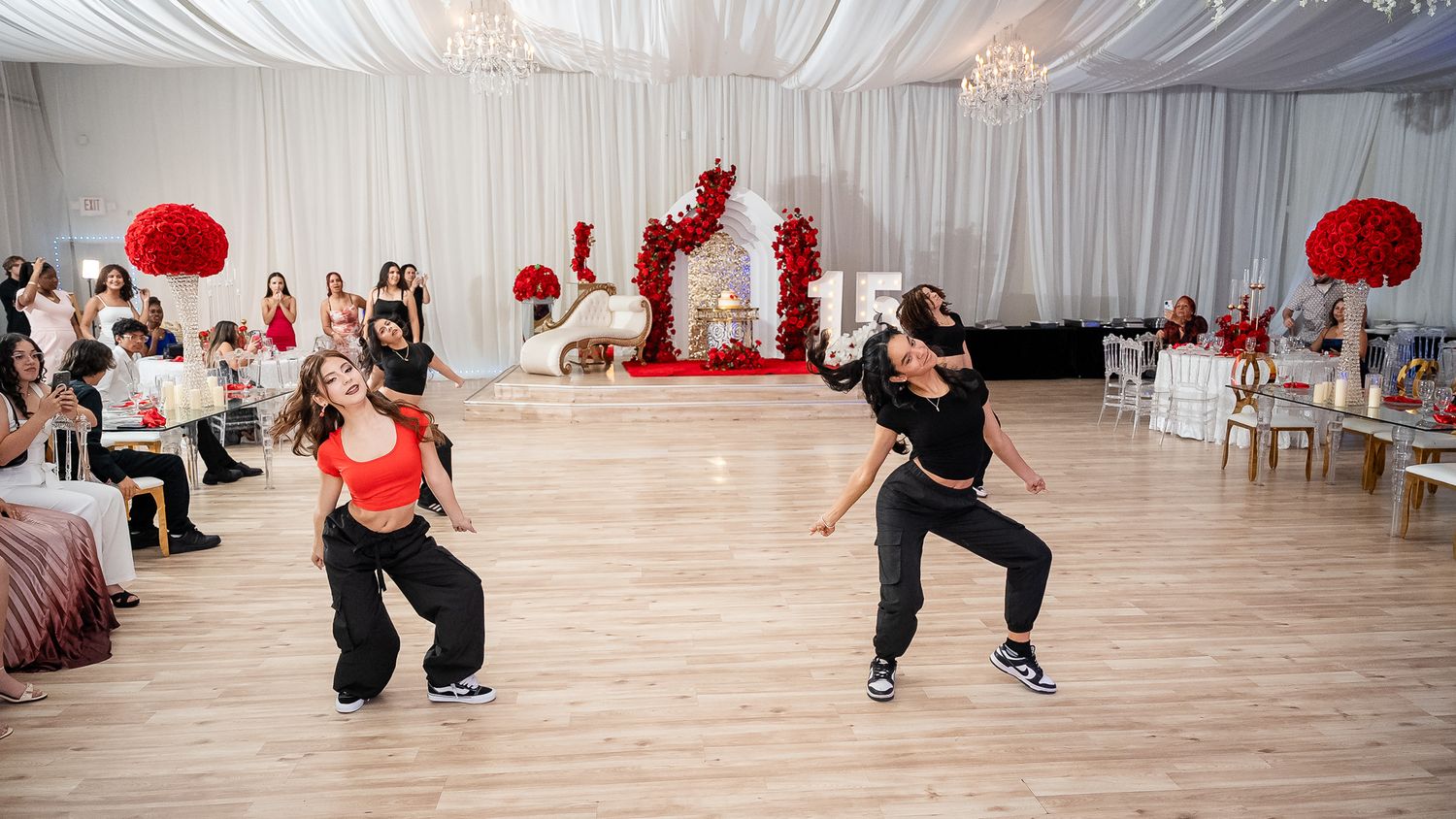 Two dancers perform at a quinceañera reception in a white draped ballroom with red floral decorations.
