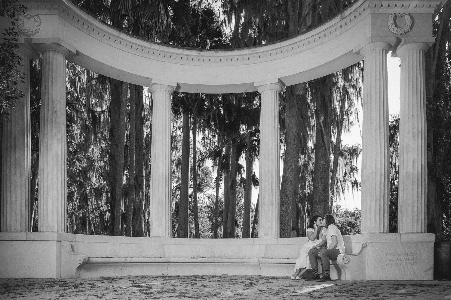 Black and white photo of a couple sitting together inside a curved colonnade surrounded by tall trees.